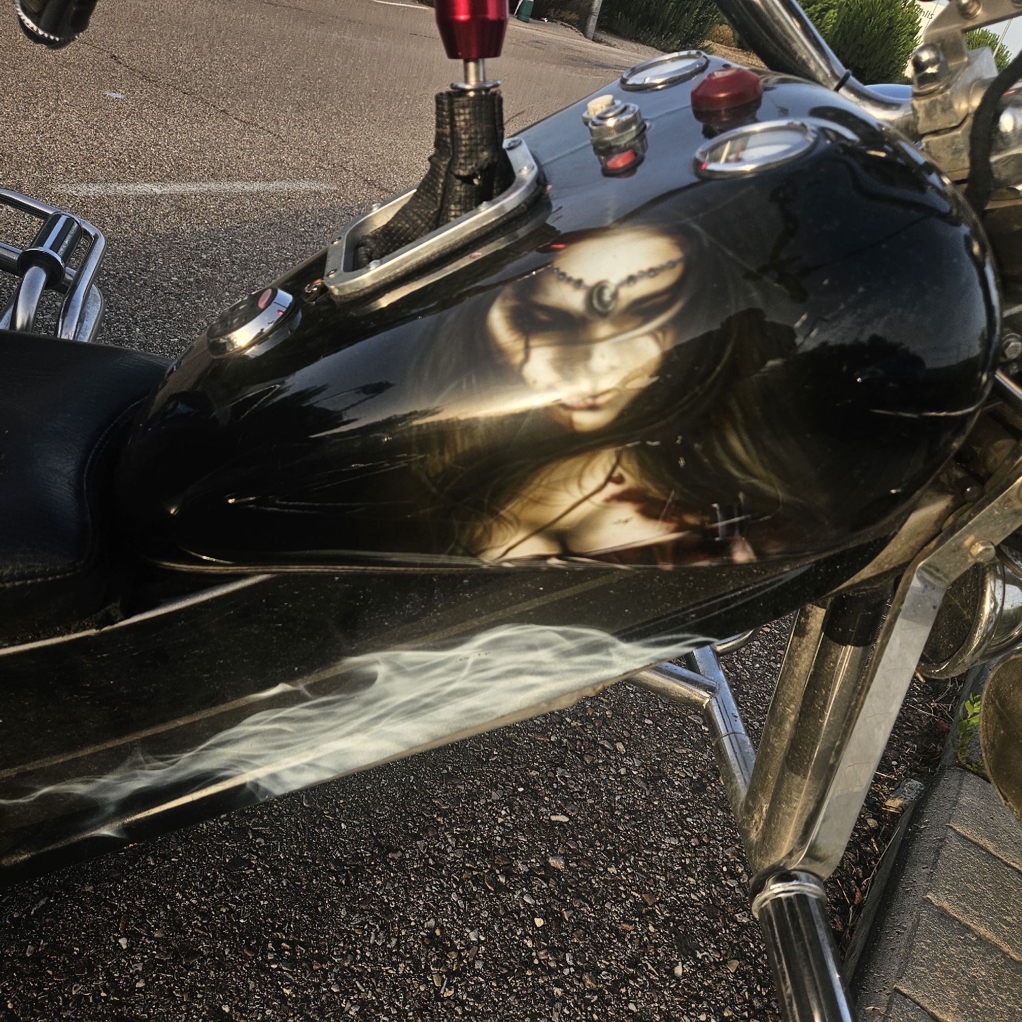 This is a close-up, eye-level shot of the fuel tank of a black motorcycle parked on an asphalt road. The motorcycle's fuel tank features a custom airbrushed design of a woman's face with dark hair, a jeweled headdress, and possibly blood splatters on her face and neck. The design is reflective and distorted due to the curved surface of the tank. White smoky graphics are also visible on the lower part of the tank.
In the background, the asphalt road surface is visible, marked with a faint white line. To the left and top of the frame, chrome handlebars and a red gear shifter with a black fabric boot are visible. The surrounding environment appears to be outdoors, with some greenery visible in the upper right corner. The image captures a detail of the motorcycle, focusing on its decorative elements and components. There are no people or actions depicted in the image. The location is the city of Illescas, Spain.