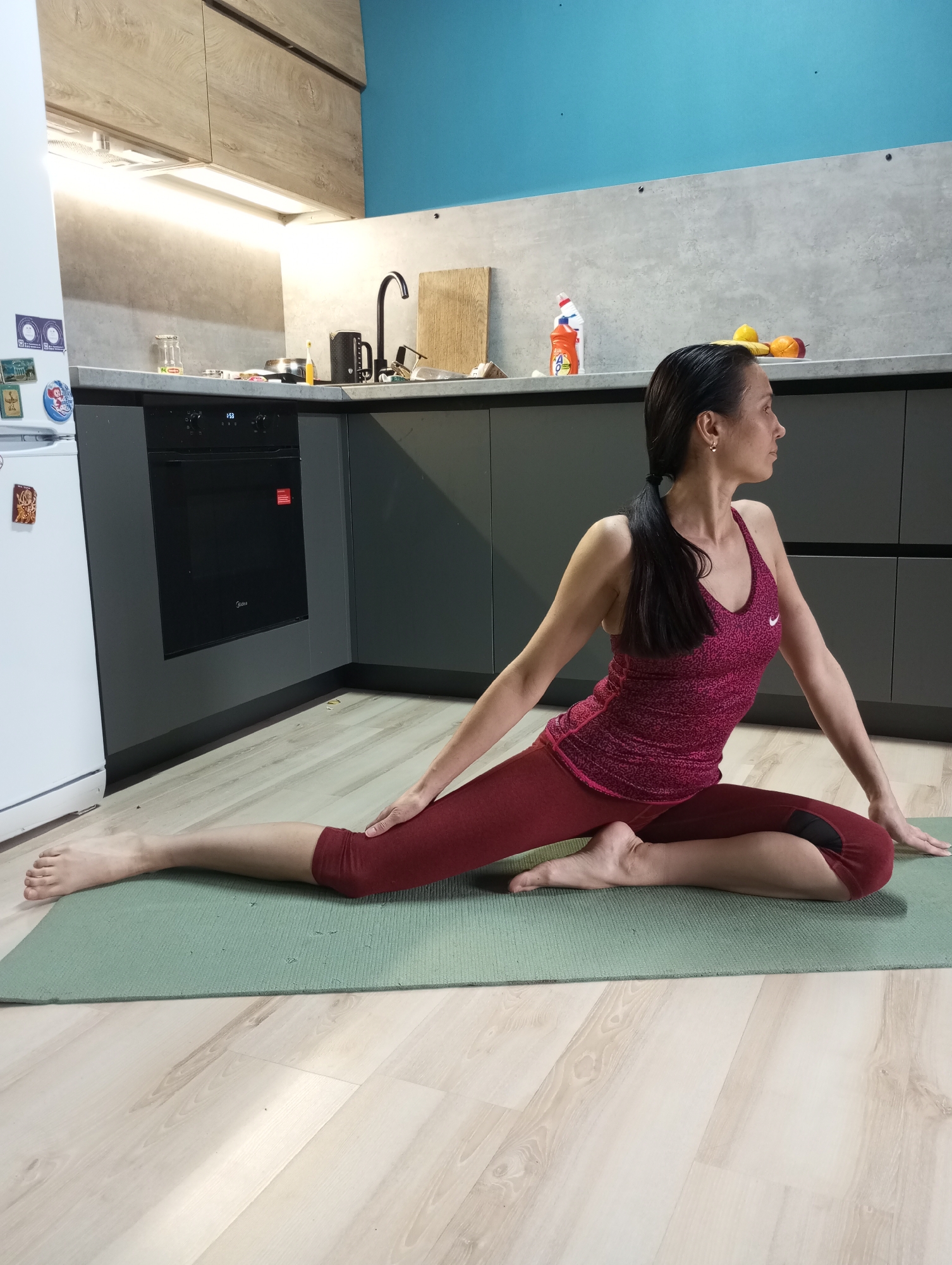 A woman is performing a yoga pose on a green yoga mat indoors. She is wearing a maroon tank top and maroon leggings. Her dark hair is pulled back into a ponytail. The room appears to be a kitchen, with gray cabinets, a black oven, and a white refrigerator visible. A blue wall is visible above the cabinets. The kitchen also contains a black faucet and other kitchen items. The floor is made of light-colored wood. This appears to be a residential setting. There is no information about the city of Pervomaysky, Russia, present in the image.