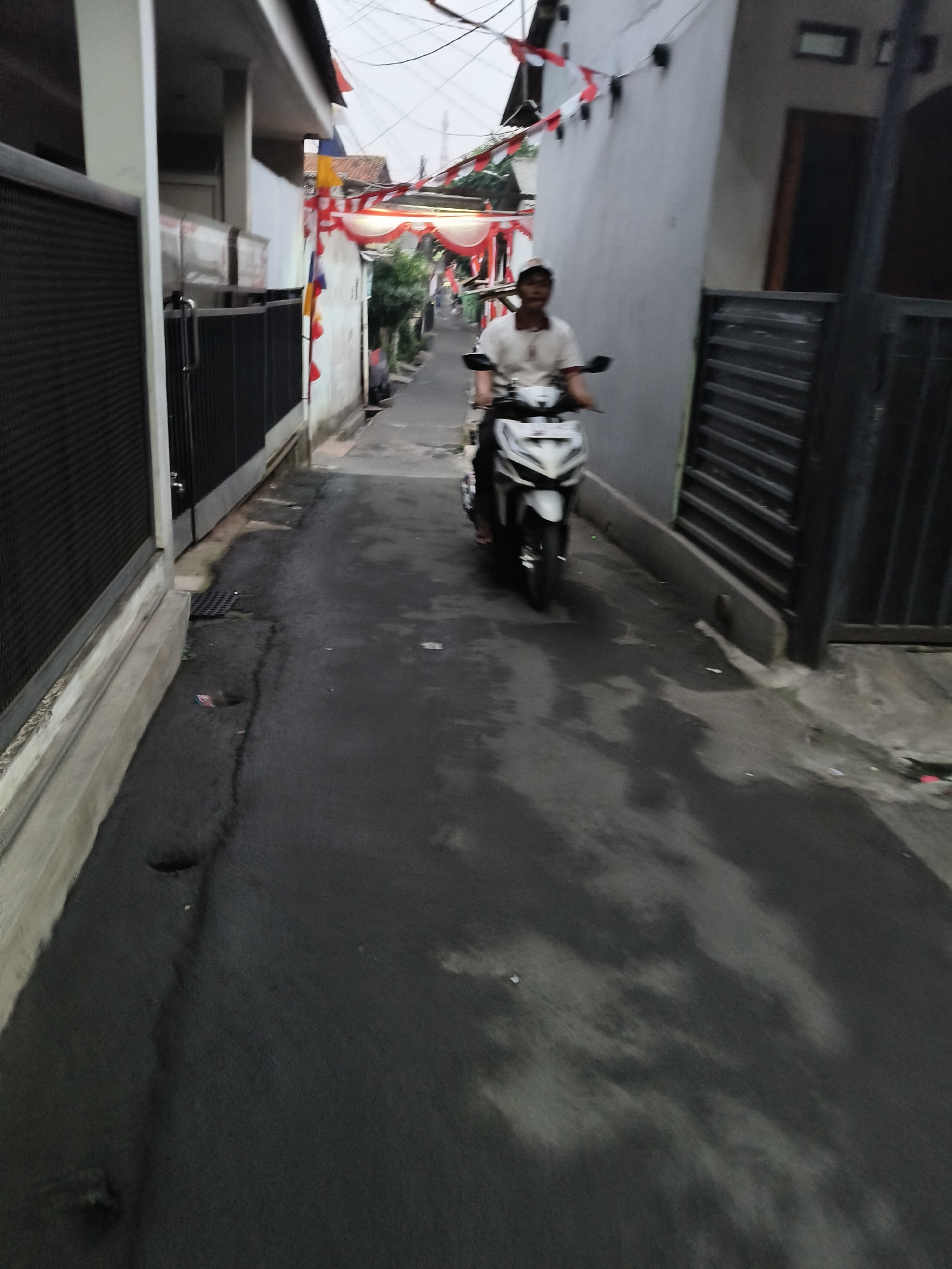 A person is riding a white motorcycle down a narrow alleyway in South Tangerang, Indonesia. The alley is lined with buildings, some with red and white flags draped across them, suggesting a celebration or national holiday. The road surface is wet and dark, with visible puddles and cracks. The person on the motorcycle is wearing a light-colored polo shirt and a cap. The lighting suggests it is either early morning or late afternoon.