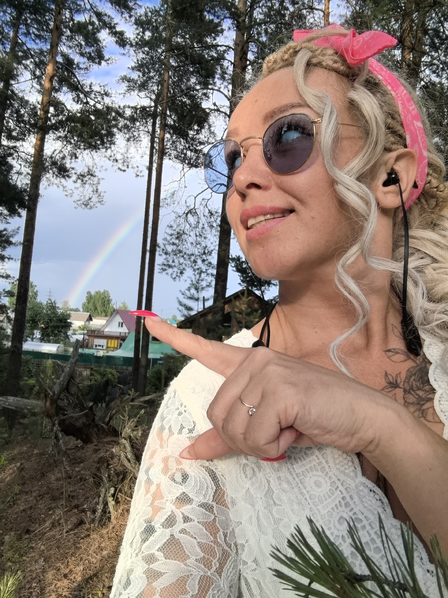 A woman with blonde braided hair and a pink bandana is standing outdoors, possibly in the city of Noviy, Russia. She is wearing sunglasses and a white lace top. She is smiling and pointing towards a rainbow in the sky, which is visible behind some trees and houses. She has a tattoo on her chest and is wearing earbuds. The environment appears to be a wooded area with some residential buildings in the background. The lighting suggests it may be daytime, possibly after a rain shower, given the presence of a rainbow.