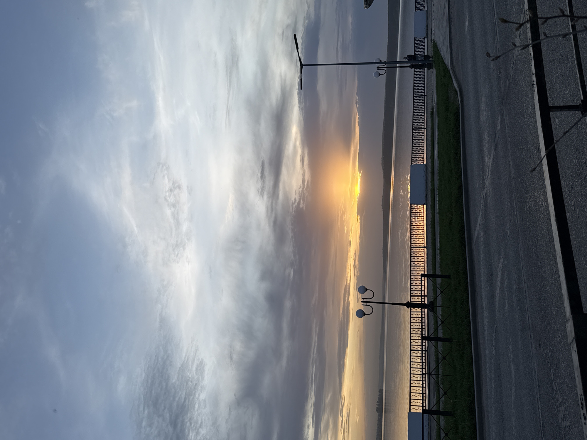 The image depicts a sunset scene in Votkinsk, Russia. The sky is filled with a mix of blue and grey clouds, with a bright orange and yellow sunset visible near the horizon. Below the sky, there is a body of water, likely the Votkinsk Reservoir, reflecting the colors of the sunset. Along the edge of the water, there is a paved embankment or promenade with a black metal fence. Street lamps are visible along the promenade. A road runs alongside the promenade, with some greenery separating the road from the fence. A tree branch is visible along the right edge of the image.