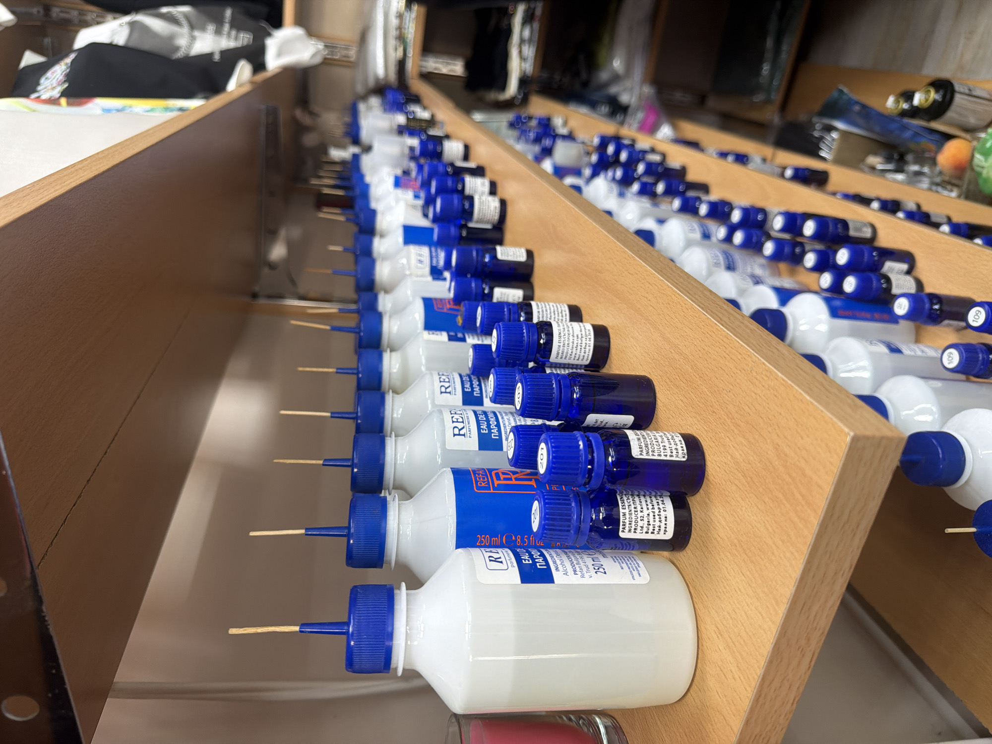 A retail establishment in Svilengrad, Bulgaria, displays shelves stocked with numerous bottles of perfume essences and alcohol bases. The foreground showcases a shelf with various sizes of white plastic bottles, some containing milky-white liquid, capped with blue plastic tops and fitted with wooden wicks. Adjacent to these are rows of small, dark blue glass bottles, also with blue caps and labels, containing perfume essences. These smaller bottles are densely packed on the shelf. The background reveals additional shelves filled with more bottles and some stacked clothing or fabric items on a higher shelf. The environment appears to be an organized retail space.