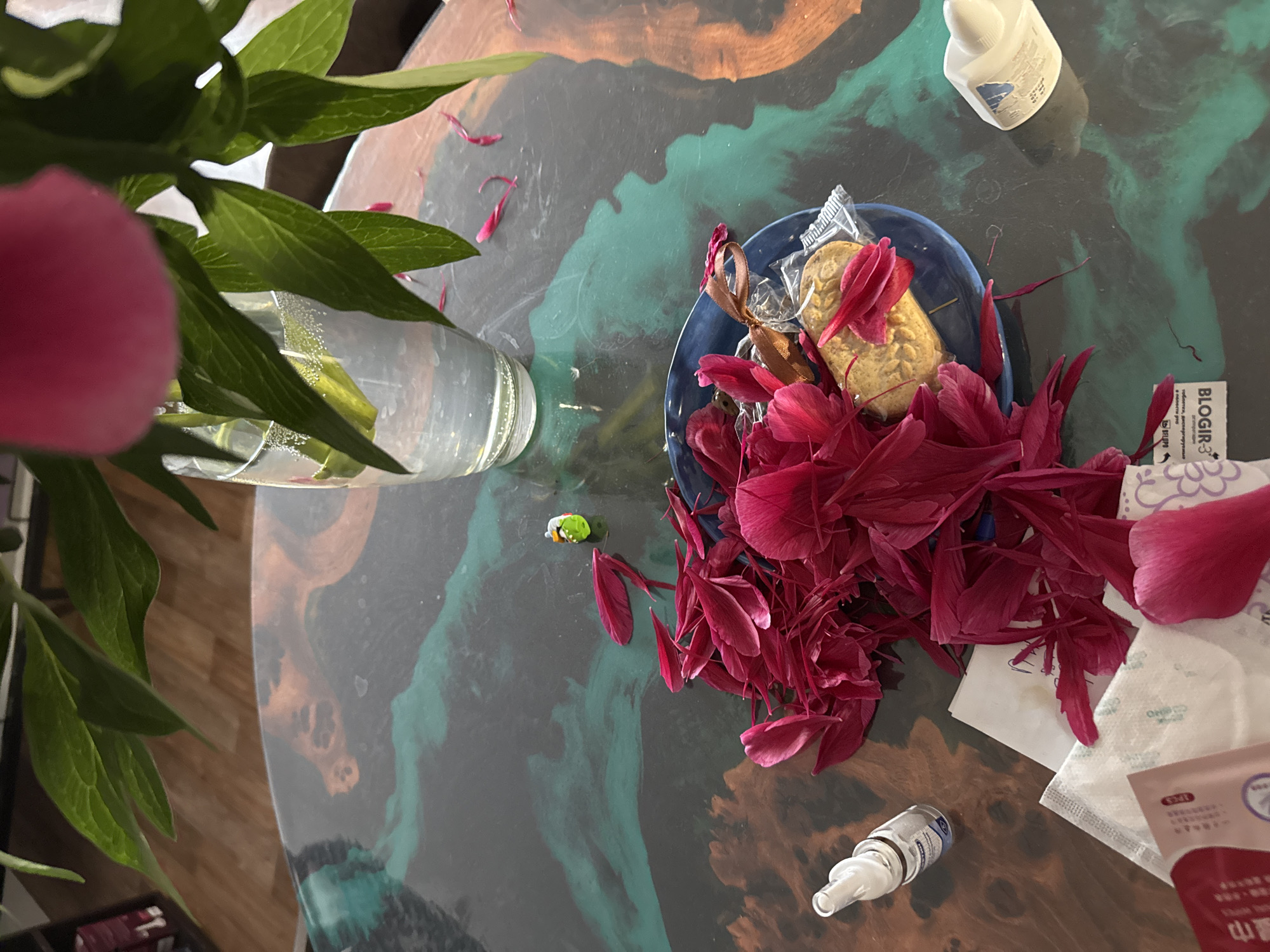 An overhead shot captures a domestic scene on a patterned, dark table, likely made of resin with swirling dark green, brown, and black tones.
In the upper left, a clear glass vase holds green foliage, appearing to be stems and leaves from a peony plant. A blurred pink flower or bud from the same plant is visible in the extreme foreground on the left. Scattered crimson peony petals adorn the table surface, with a larger concentration of these petals inside a small blue bowl located centrally on the right.
Inside the blue bowl, amongst the petals, are two individually wrapped cookies, one tied with a brown ribbon, suggesting a snack. Various small items are also present on the table. In the upper right corner, a small white dropper bottle with a white cap is visible. Next to the bowl, a white packet with "BLOGIR-3" (and likely "БЛОГИР-3" in Cyrillic, aligning with the Perm, Russia context) is partially covered by petals. Another white, patterned packet and a red packet with Asian characters (including "山雲") are situated towards the bottom right. A clear glass bottle with a dropper or nozzle is also on the table near the bottom right.
The scene suggests a casual indoor setting, possibly after a peony has bloomed and shed its petals, combining elements of natural beauty with personal items and snacks.