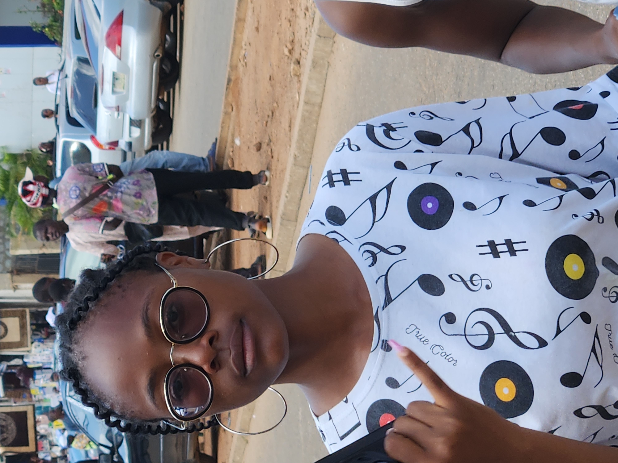 A young woman with braided hair and large hoop earrings, wearing sunglasses and a white t-shirt with a musical note pattern, is the main subject of the image. She is in a shallow, upright crop of a person. She is looking into the camera, and her right index finger is extended, pointing towards a "True Color" graphic on her shirt. The background is blurred, but shows a sandy ground, some vehicles, and other people, suggesting an outdoor setting, possibly a market or a street. The image is taken during the daytime, with bright lighting. The woman's expression appears neutral.