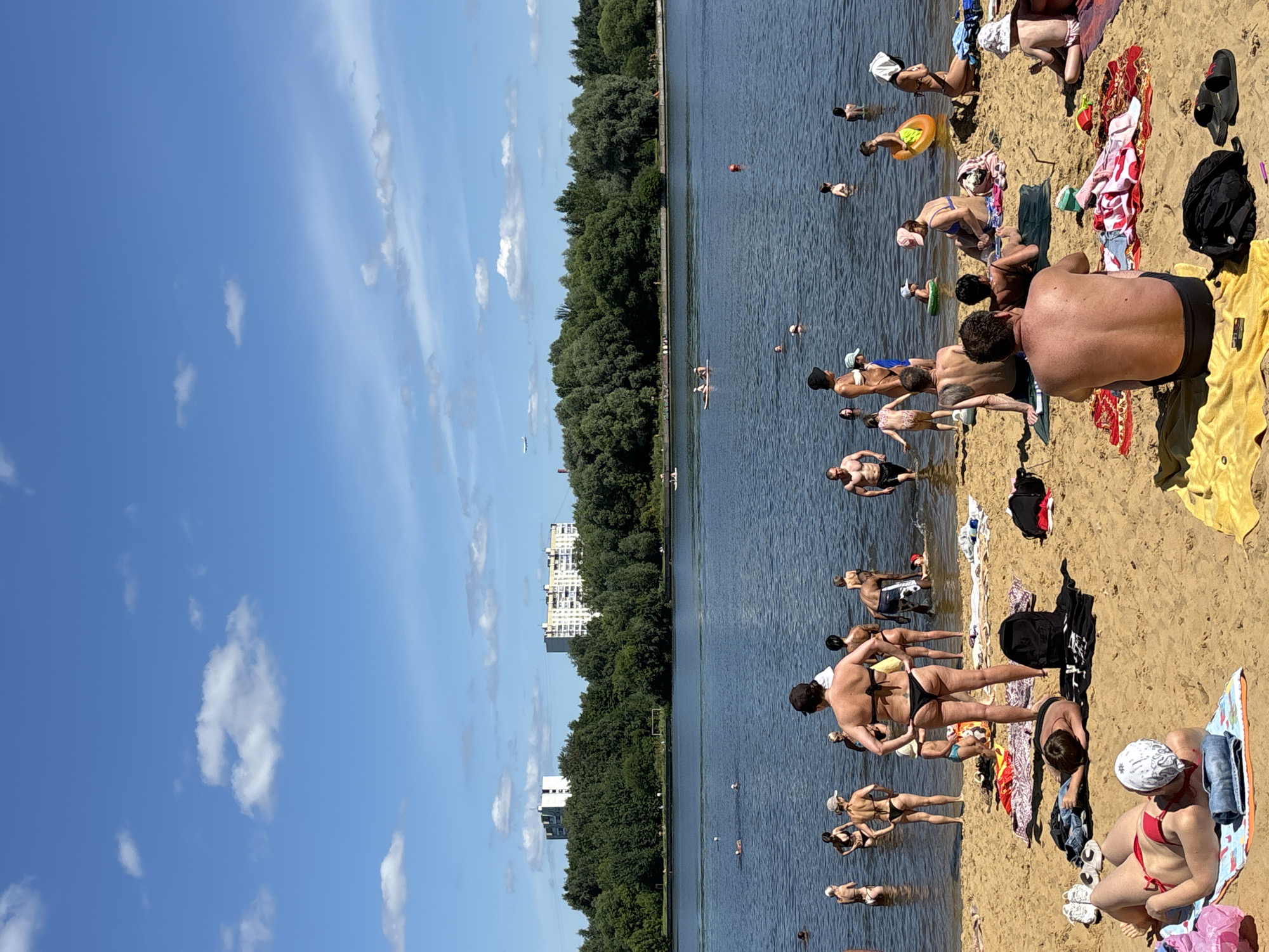 In the city of Dolgoprudny, Russia, a sandy beach on a sunny day is filled with people enjoying the water and sun. The scene captures a lively atmosphere with individuals engaged in various beach activities.
In the foreground, several people are relaxing on the sand. A woman in a red bikini and a white headscarf is lying on a towel, looking at the camera. Next to her, a person is lying face down on a towel, wearing a black bikini. A man with his back to the camera is sitting on a yellow towel, wearing black swim trunks. Another person is sitting next to him on a dark green towel.
Further back, numerous people are wading in the shallow water or standing at the water's edge. Some are dressed in swimwear, while others are partially submerged. Groups of people are scattered across the beach, with towels, bags, and inflatable toys visible.
The background features a calm body of water, likely a reservoir or lake, reflecting the blue sky dotted with white clouds. Across the water, a dense line of lush green trees creates a natural barrier. Beyond the trees, several tall, multi-story apartment buildings are visible, indicating the presence of a residential area. The sky is clear blue with a few scattered clouds and an airplane flying in the distance. The overall impression is a typical summer day at a popular urban beach.