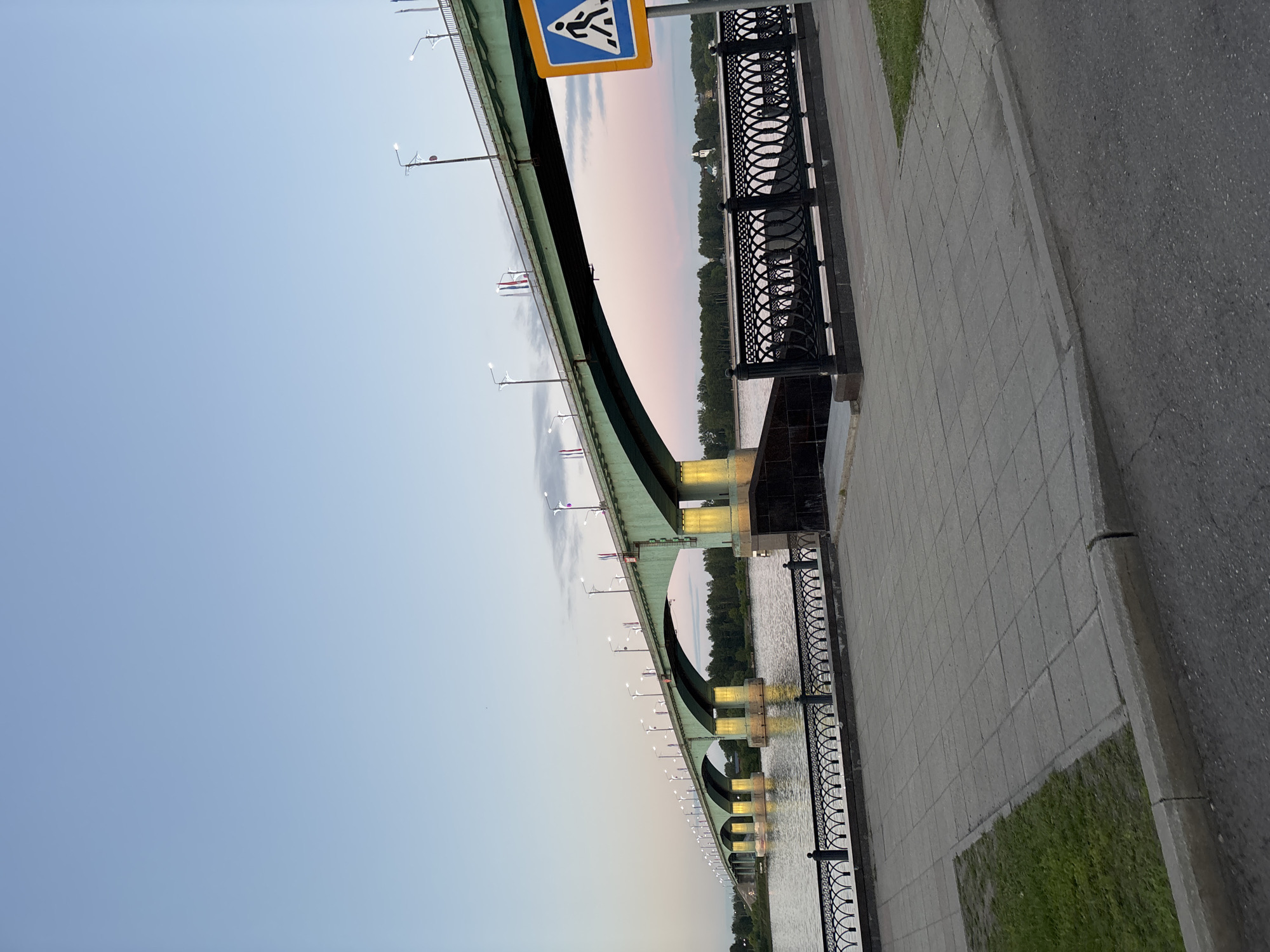 The image depicts a bridge and a pedestrian walkway in Rusanovo, Russia. The bridge has a distinctive arched design, with support structures and lighting fixtures visible along its length. A pedestrian crossing sign is positioned near the bridge. The walkway features a black, ornate railing along the water's edge. The surrounding environment includes water, trees lining the opposite bank, and a clear sky. Adjacent to the walkway is a paved area and a strip of grass. There are no people visible in the image.