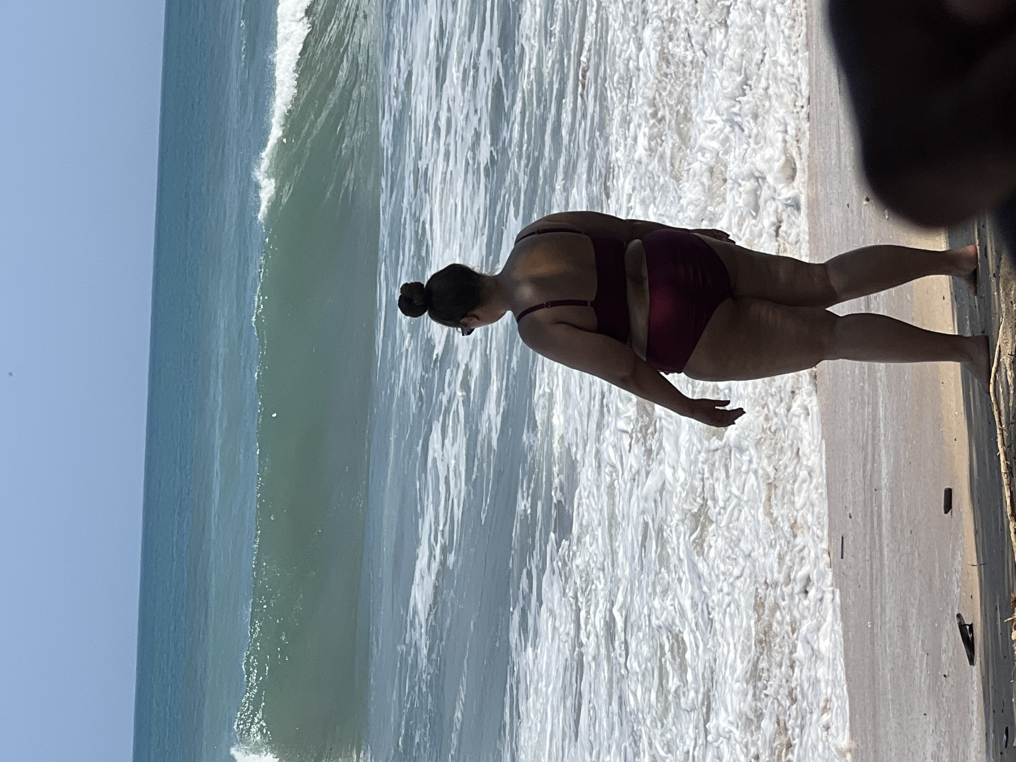 A woman wearing a maroon bikini stands on a beach in Kuta, Indonesia, facing the ocean. The water is a turquoise blue with a moderate wave breaking in the distance. The sand is light brown, with some small pieces of debris scattered on it. The sky is a clear, pale blue. The woman has her hair pulled up in a bun and is wearing sunglasses. The image is taken from a slightly low angle, emphasizing the size of the waves.