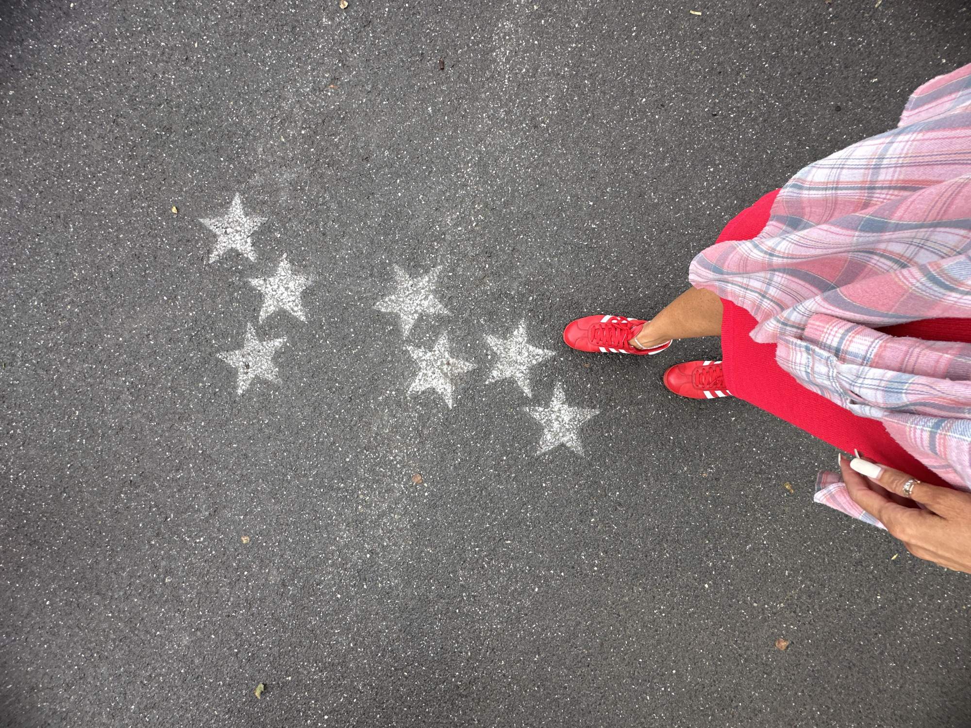 The image depicts a top-down view of asphalt pavement with several white, stenciled stars. A person's legs and lower torso are visible, wearing bright red sneakers with white stripes and a vibrant red skirt. A plaid shirt, predominantly in shades of pink and blue, is draped over the person's lower body. The person's left hand, with long white-nailed fingers and a ring on the ring finger, is in the foreground, resting near the plaid shirt. The environment appears to be an outdoor urban setting. There is no discernible information or visual cues in the image to suggest the location is the city of Tyumen, Russia.
