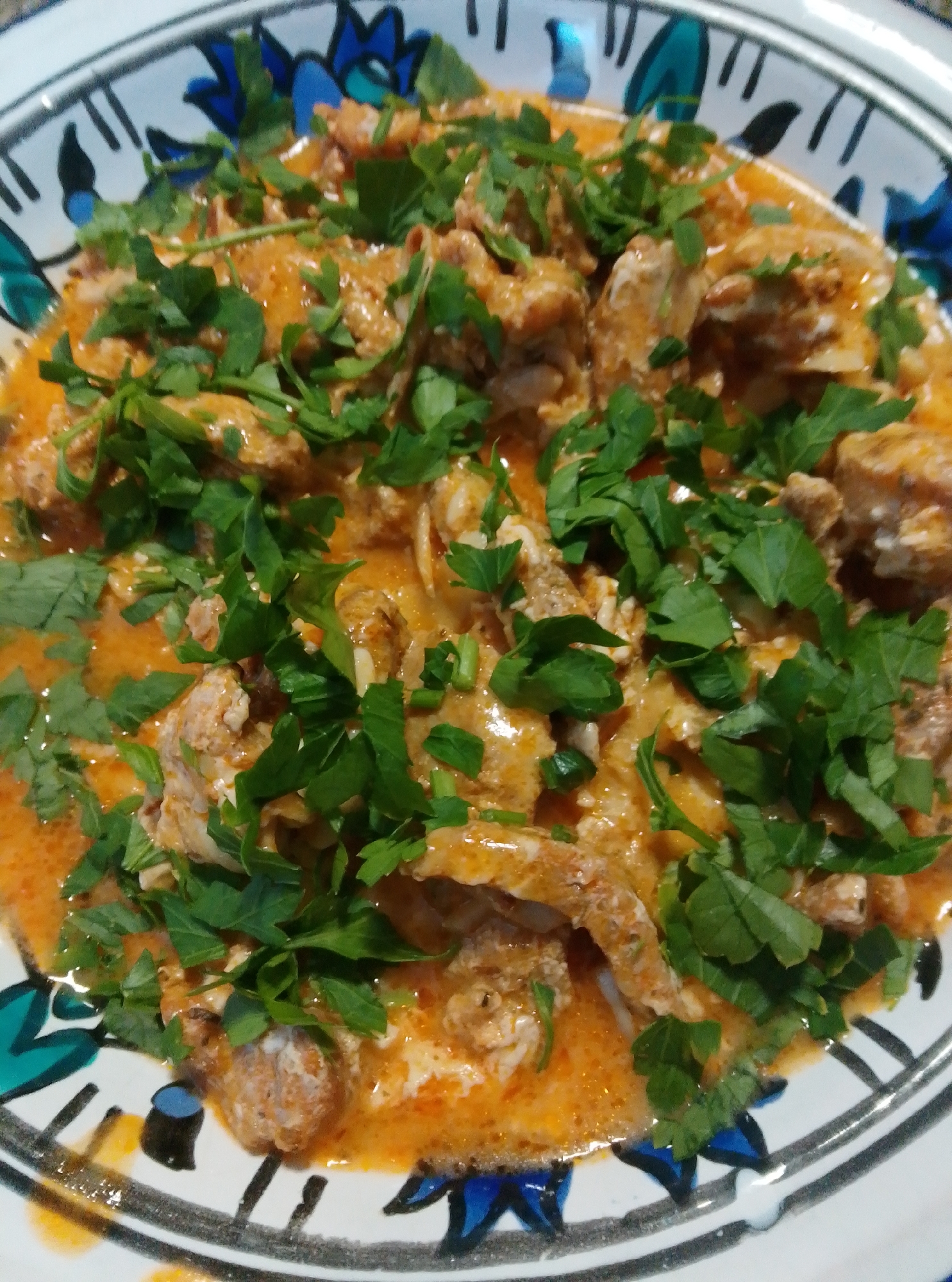 A close-up, overhead shot captures a bowl filled with a creamy, orange-hued stew, likely a seafood or chicken dish. The stew is generously garnished with chopped fresh parsley, providing a vibrant green contrast to the warm tones of the food. The bowl itself is white with a decorative rim featuring abstract blue and black designs. The lighting suggests an indoor setting, possibly a home kitchen or restaurant. No people or specific location in Wedel, Germany, are visible in the image.