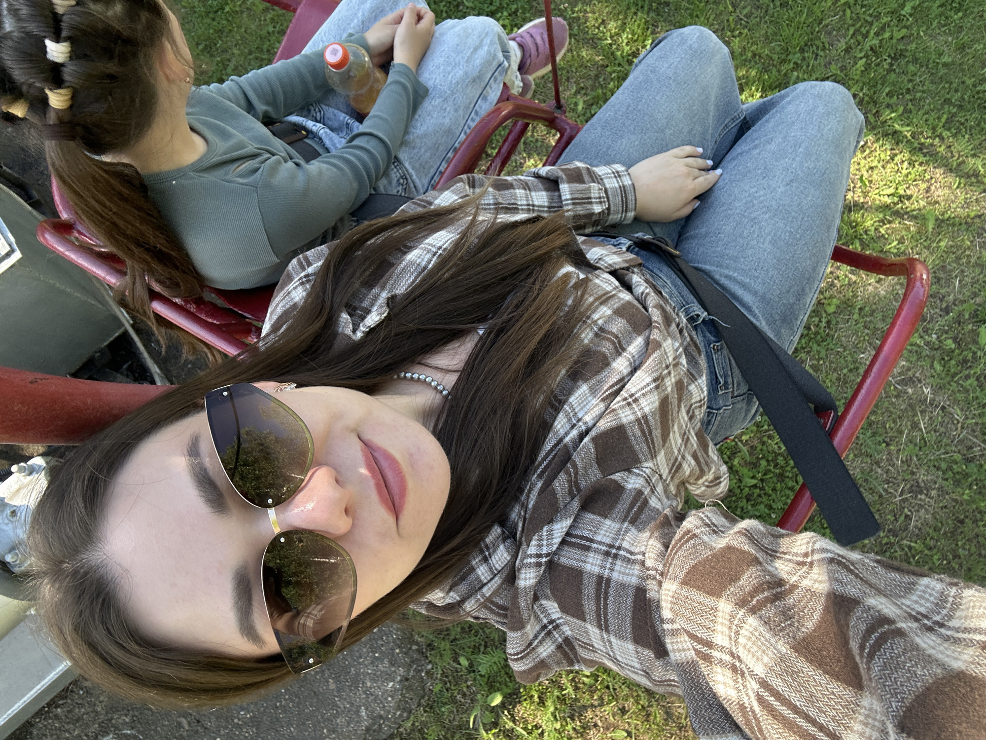 Two young women are seated on a red metal chair, likely a ski lift, outdoors in Chaikovsky, Russia. The foreground woman wears sunglasses, a brown plaid shirt, jeans, and a pearl necklace. Her hair is brown and straight. The background woman, only partially visible, wears a green long-sleeved shirt, jeans, and has her hair in a ponytail with multiple hair ties. A plastic water bottle is near her. The background shows green grass.