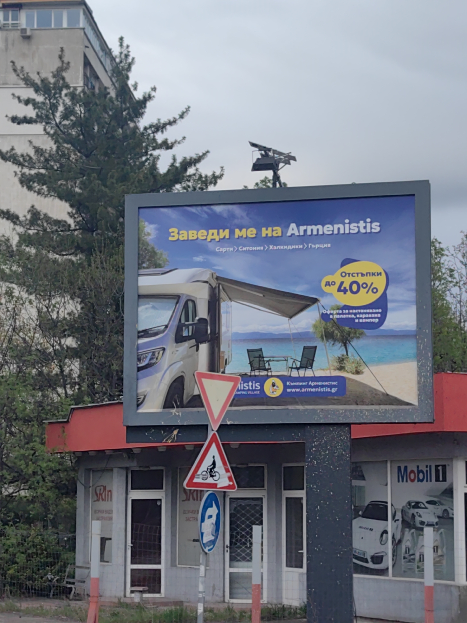 The image captures a street scene in Sofia, Bulgaria, featuring a prominent billboard advertising a travel destination. The billboard, centrally located, displays a large camper van parked on a sandy beach next to the sea, with an awning extended and two chairs and a table set up. The text on the billboard is in Bulgarian, reading "Заведи ме на Armenistis" (Take me to Armenistis), followed by "Сарти > Ситония > Халкидики > Гърция" (Sarti > Sithonia > Halkidiki > Greece). A yellow oval highlights a discount of "Отстъпки до 40%" (Discounts up to 40%), with an offer for "настаняване в палатка, каравана и кемпер" (accommodation in a tent, caravan, and camper). The website "www.armenistis.gr" is also visible.
Below the billboard is a single-story commercial building with a red roof. On its left, a shop window shows the text "Rin" and partially visible "ЗАСТРА" (possibly for insurance, "Застраховане"). On the right, another shop front displays "Mobil 1" branding and images of white Porsche cars, suggesting an automotive-related business.
In the foreground, three road signs are visible on a pole: an inverted red-bordered triangle indicating "Give Way," a circular sign with a red border and a bicycle, likely meaning "No Cycling," and a blue circular sign with a white arrow pointing right, indicating a "Mandatory Right Turn" or "Direction to be followed."
To the left of the billboard, a tall, multi-story building with windows is partially visible, alongside a large, dense evergreen tree. The sky is overcast, suggesting daytime. Above the billboard, a structure with what appears to be solar panels or a lighting rig is mounted on a pole.