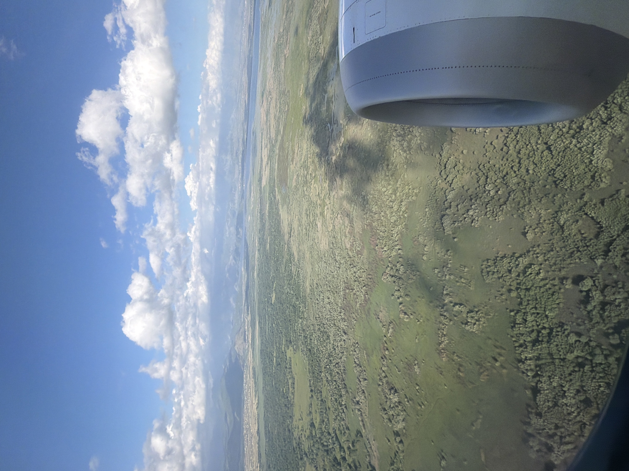 An oblique aerial view from an aircraft window captures a landscape of green vegetation, likely forests and meadows, interspersed with possibly small settlements. A part of the airplane’s engine is visible in the upper right corner of the frame. The sky, with fluffy white clouds, occupies the left portion of the image. A distant body of water is visible near the horizon. The scene suggests a rural or sparsely populated area, possibly somewhere near Gostilj, Montenegro.