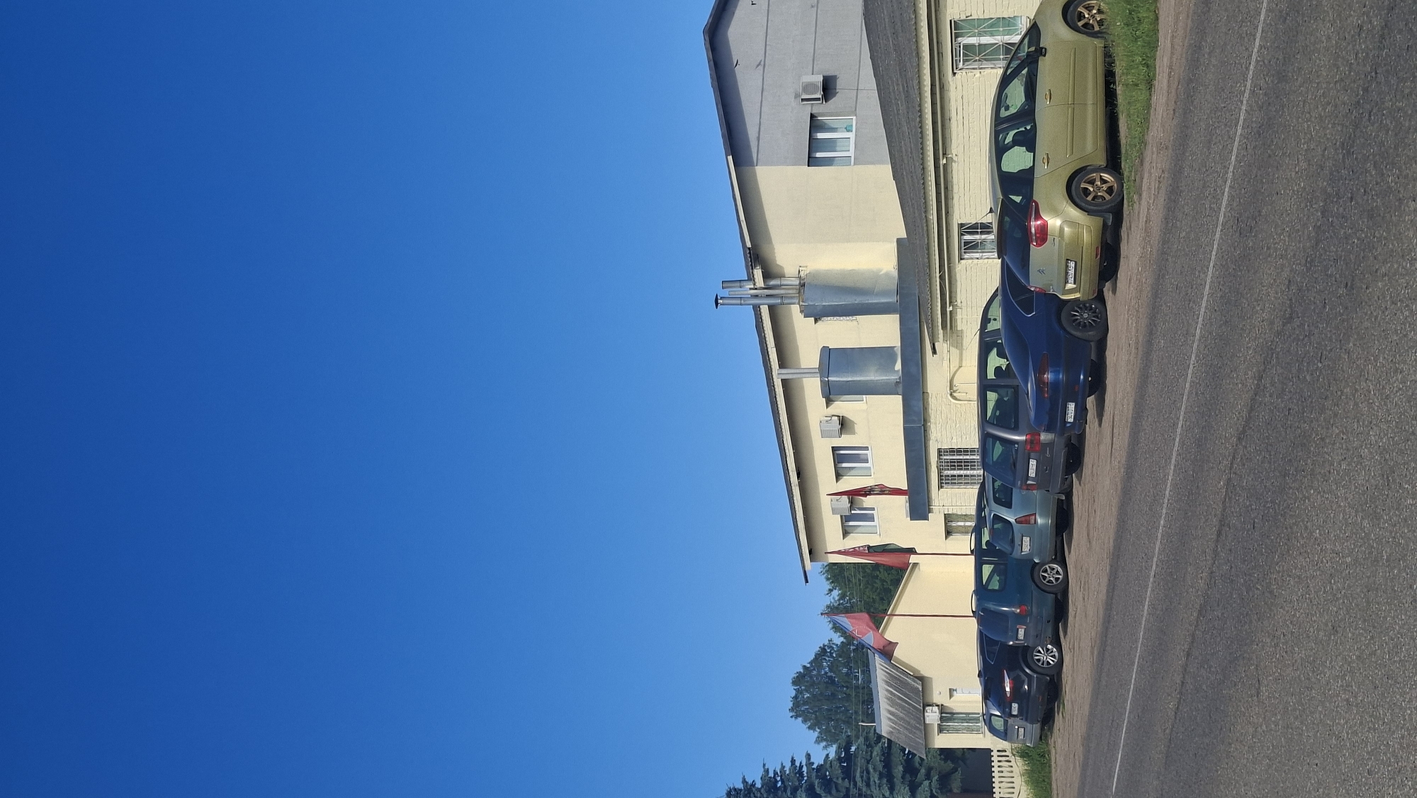 This is a photograph of a building in Забалоцкі сельскі Савет, Belarus, on a clear, sunny day. The building is a light yellow two-story structure with a gray roof and several windows. Two prominent flags are flying from poles in front of the building. Multiple cars are parked in a row along the front of the building, adjacent to a paved road. The environment appears to be a rural or suburban area, with trees visible in the background.