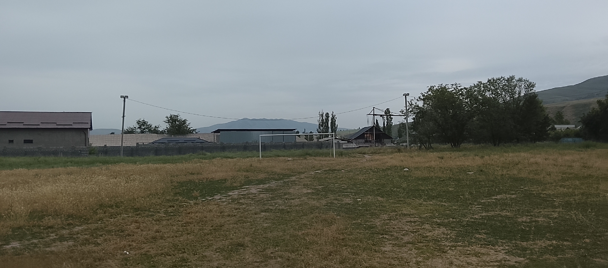 The image depicts an outdoor scene in an undeveloped area. In the foreground, a dry, patchy grass field is visible, with a faint path marking across it. A white soccer goal stands in the middle ground. Behind the field, a low concrete wall separates it from a cluster of buildings. To the left, a rectangular building with a dark, sloping roof and small windows is present. Further to the right, several other structures with tiled roofs are visible. Beyond the buildings, a line of trees runs horizontally, with some taller, slender trees standing out. In the background, a broad, flat-topped mountain range is silhouetted against an overcast sky. The overall impression is one of a rural or semi-rural location.