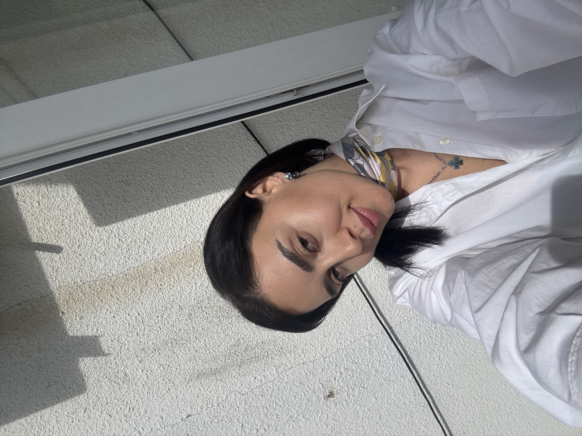 The image shows a woman in a white shirt, wearing a colorful neck scarf and a cross necklace. She is standing against a white, rough-textured wall. The lighting suggests it is daytime. A window frame is visible above her.
Without additional context, identifying the exact location in Tbilisi is not possible.