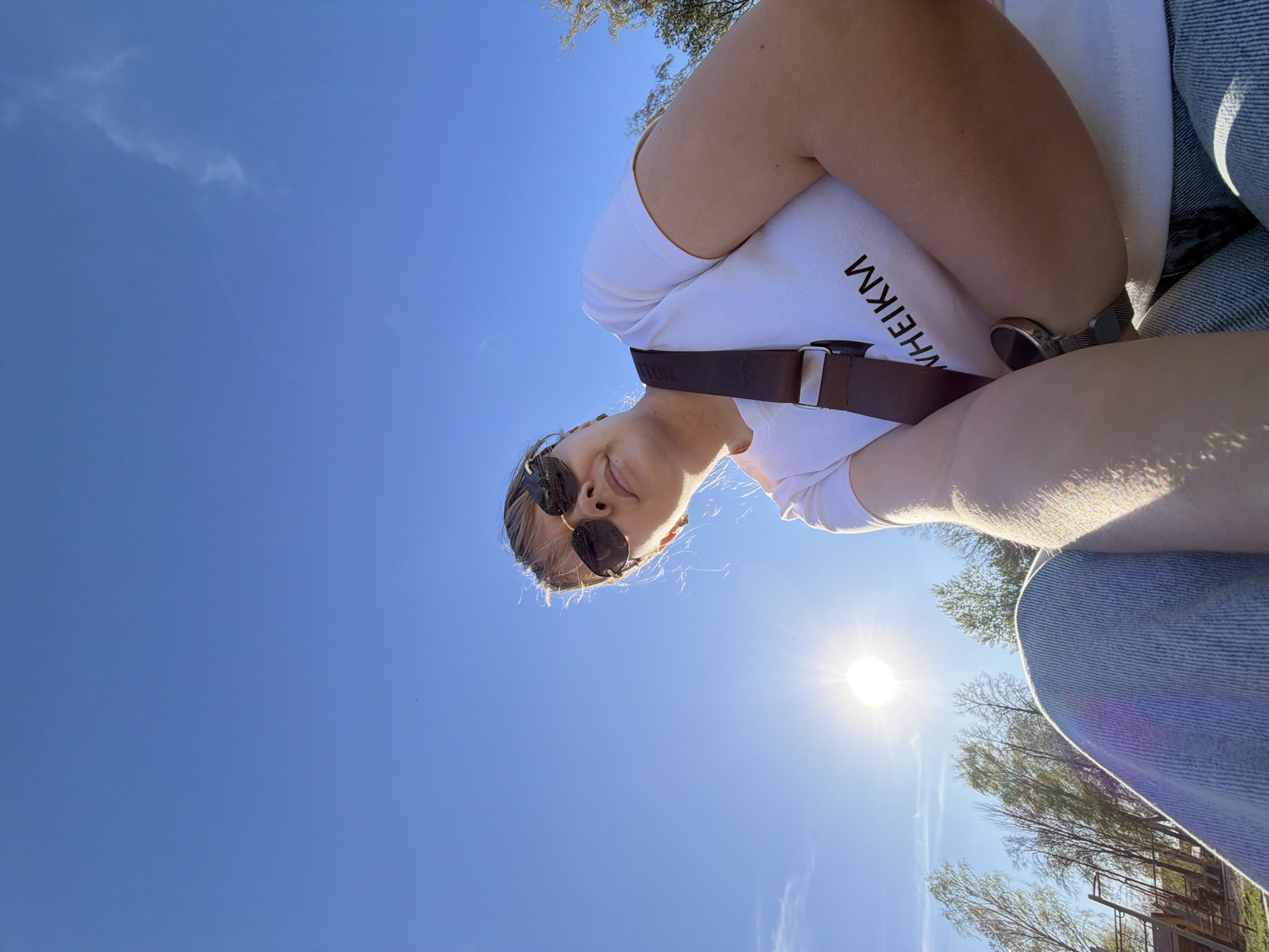 A selfie taken at a low angle shows a person with blond hair, wearing sunglasses, a white t-shirt with the word "WHEIM" printed on it, and blue jeans. The person is wearing a dark strap, possibly for a bag, across their chest. Part of a watch is visible on the person's left wrist. The background features a bright blue sky with some cirrus clouds. The sun is shining brightly, causing some glare. Bare trees are visible in the distance. The location appears to be outdoors in daylight, although specific details connecting it to Chaikovsky, Russia, are not discernable from the image alone.