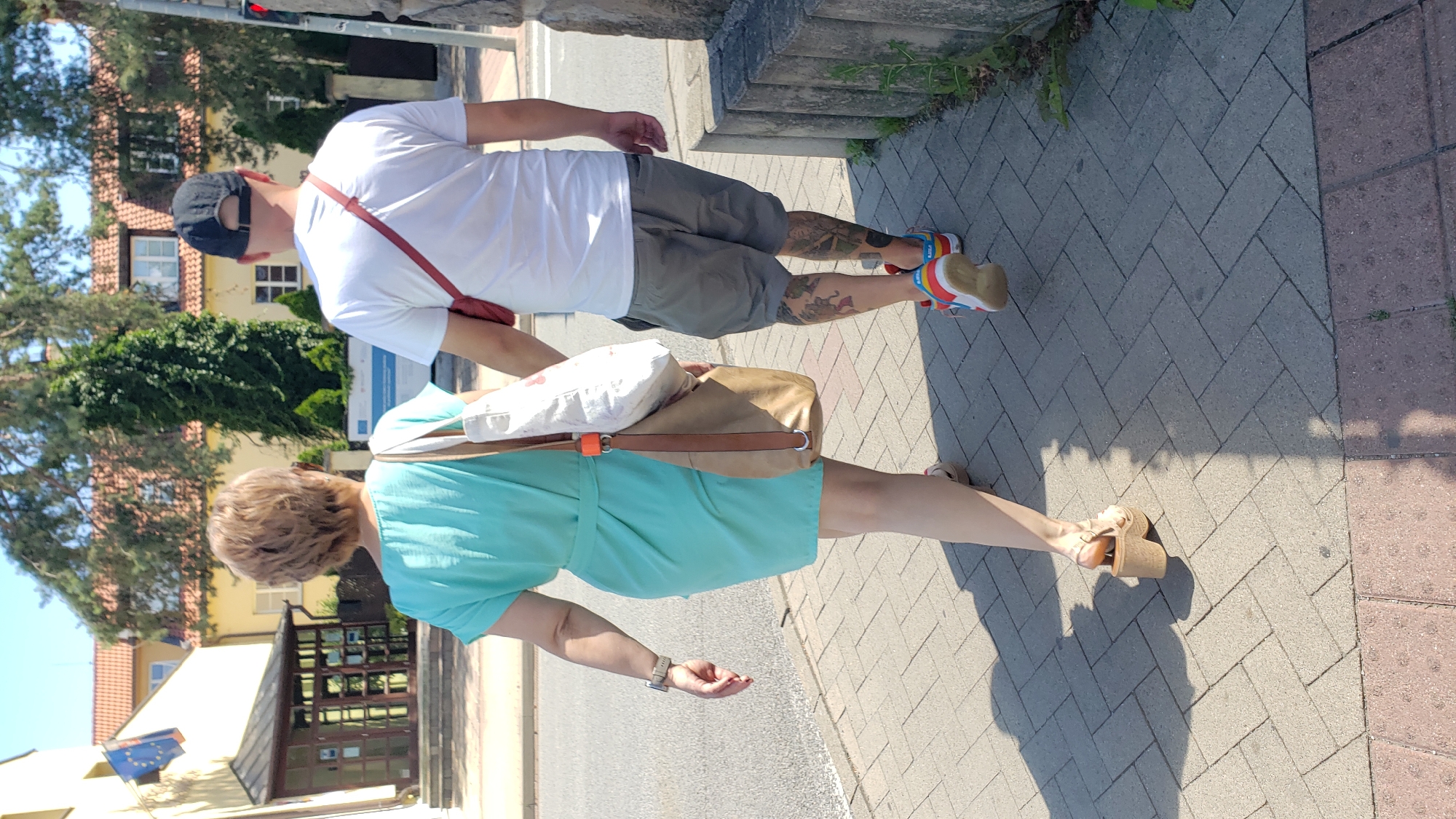 Two individuals, a woman and a man, are walking away from the camera on a paved sidewalk. The woman, on the left, wears a turquoise dress and beige platform sandals. She carries a beige shoulder bag with a white item inside. The man, on the right, wears a white t-shirt, grey shorts, a black baseball cap, and colorful sneakers. He has tattoos on his legs. A red strap is visible across his chest. They are walking in the direction of a building with a yellow facade and a red-tiled roof. A flag is visible on the left side of the building. The environment suggests an urban setting, possibly Prešov, Slovakia, given the context. The lighting indicates a sunny day.