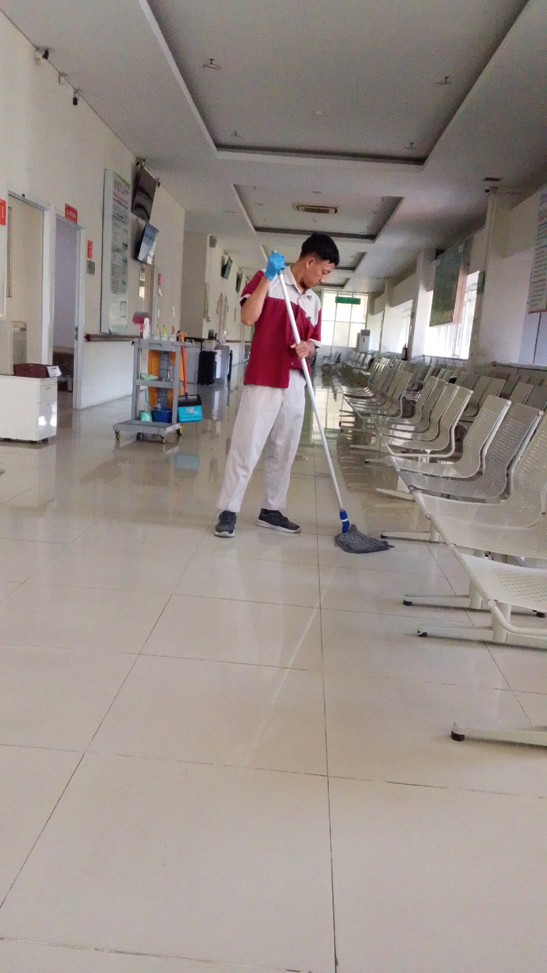 A male custodian wearing a maroon and white uniform and blue gloves is mopping a tiled floor in a brightly lit interior hallway. He is holding a mop with a gray head and a long metal pole, actively cleaning the floor. The hallway features rows of empty, light-colored metal chairs for seating on the right side. On the left side, there are doors, possibly to examination rooms, and a cleaning cart with various supplies. The ceiling has modern lighting fixtures. The overall environment appears to be a clinic or hospital waiting area, likely located in Pabelan, Indonesia.