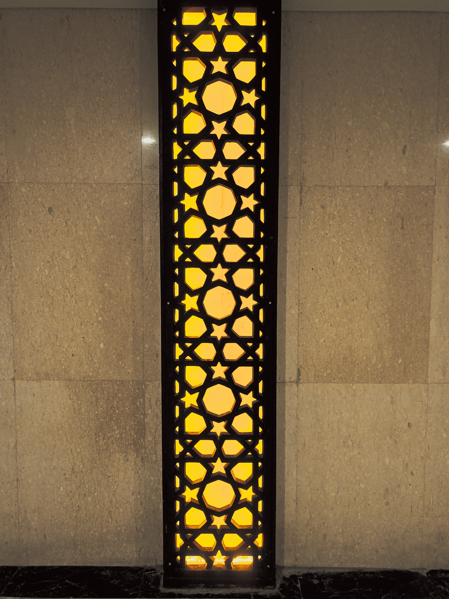 A vertical decorative panel with a geometric Islamic star pattern is set into a wall of tan-colored stone tiles. The panel is backlit with a warm, yellow light. The bottom edge of the wall has a dark-colored baseboard. The location is Banda Aceh, Indonesia.