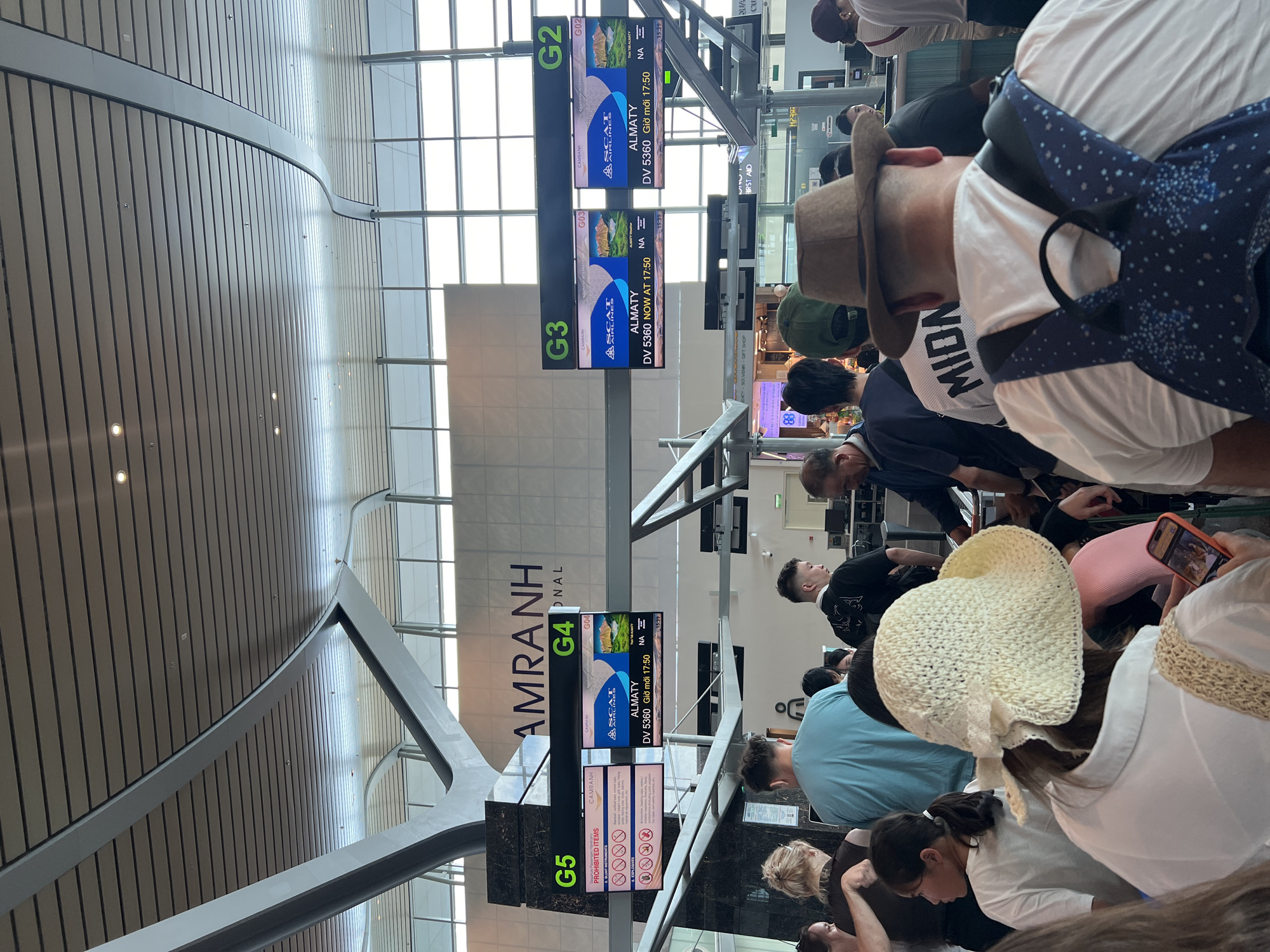 The image captures a crowded indoor scene at Cam Ranh International Airport in Bac Cam Ranh Ward, Vietnam. Numerous people, seen mostly from behind, are gathered or queuing. The setting features a modern airport terminal with a high, architecturally designed ceiling composed of wooden panels and large windows allowing natural light.
Several digital display screens are prominent, labeled G2, G3, G4, and G5, indicating gate numbers. Screens G2, G3, and G4 display flight information for "ALMATY" (Almaty, Kazakhstan) with flight number "DV 5360" operated by "SCAT AIRLINES," with a departure time of "NOW AT 17:50" or "Giờ mới 17:50" (New time 17:50). Screen G5 shows "PROHIBITED ITEMS" with corresponding warning icons. In the background, large letters spell out "AMRANH PNAL," clearly indicating "Cam Ranh International Airport." Other signs visible include "SOUVENIR GIFT SHOP" and "FIRST AID."
People in the foreground and midground wear casual attire, including hats and t-shirts, with one person wearing a hat and a shirt partially visible with the text "MIDN." Many appear to be waiting patiently, some looking at their phones. The overall atmosphere is that of a busy airport during late afternoon or early evening, given the 17:50 departure time shown on the flight screens.