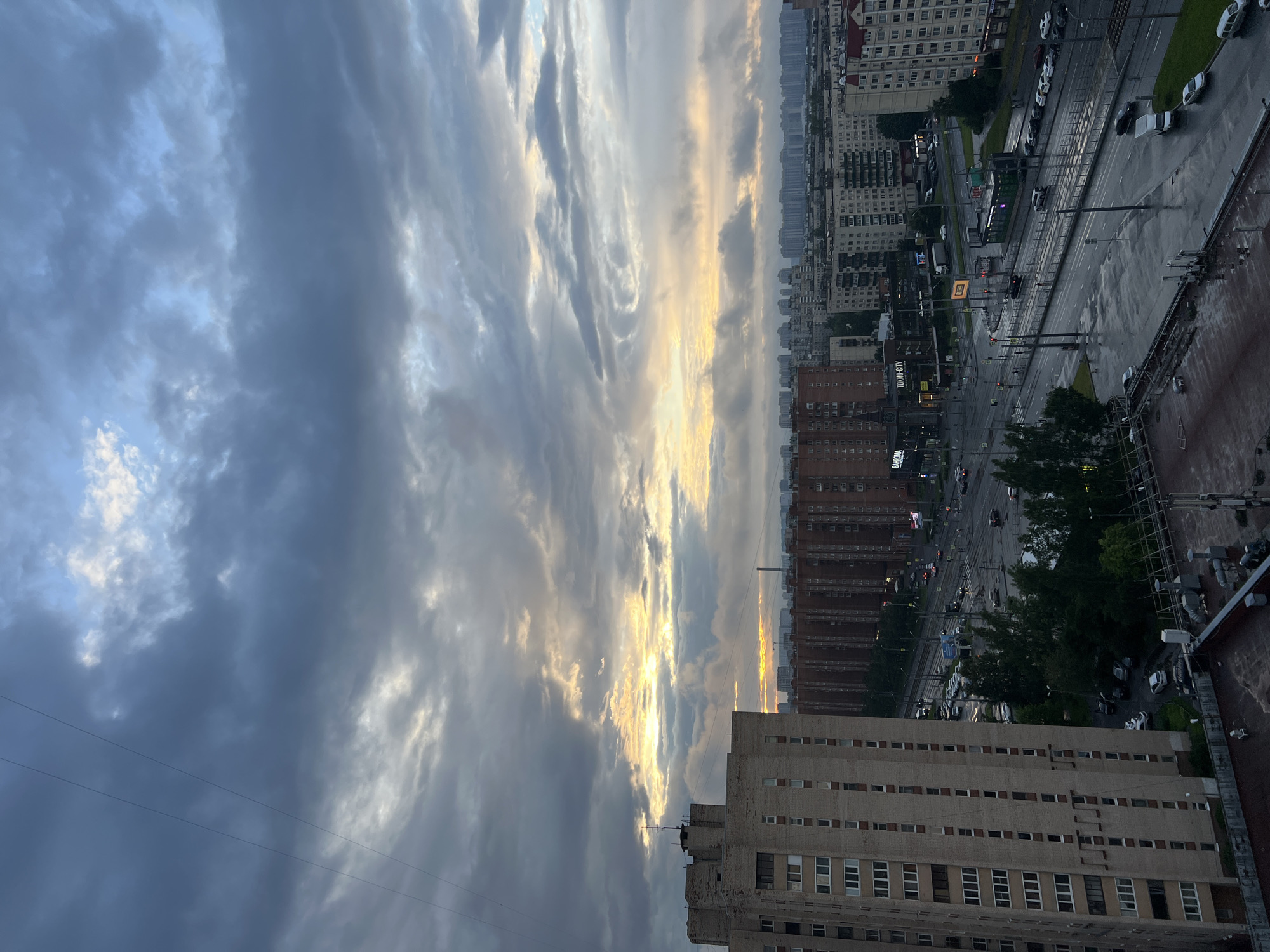 The image captures an overcast sky over a dense urban landscape in an unknown city in Russia. The sky is filled with a mix of dark gray and lighter, textured clouds, with a sliver of pale yellow light breaking through near the horizon, suggesting either sunrise or sunset. In the foreground, a tall, beige brick apartment building dominates the left side of the frame. Beyond it, a long, reddish-brown apartment complex stretches horizontally across the middle ground, punctuated by darker windows.
A wide, wet road curves through the city, with traffic lights and multiple lanes visible. Several cars are on the road, some moving, others parked along the sides. Green trees line parts of the road, creating a contrast with the surrounding concrete and brick structures. Further in the distance, a sprawling cityscape of apartment buildings extends to the horizon under the muted sky. The wetness of the road indicates recent rain. There are no clearly discernible people in the image. The overall impression is one of a residential urban area during or shortly after a period of rain, with subdued natural light.