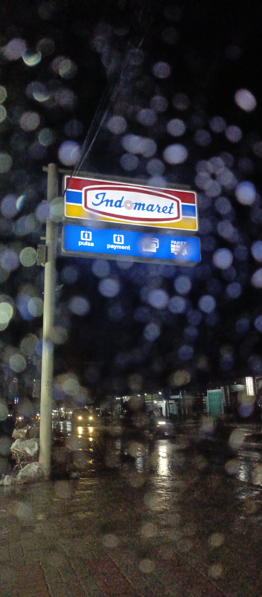 The image displays an urban night scene in Panterakyat, Indonesia, viewed through a surface covered in numerous out-of-focus water droplets, creating a distinct bokeh effect from light sources. A prominent, illuminated Indomaret convenience store sign, featuring the brand name in white text with a red outline on a blue and yellow background, is centrally positioned. Below it, a smaller blue sign displays icons and text for "pulsa" (phone credit/top-up) and "payment," with a partially visible third option, "paket" (package services). The sign is affixed to a light-colored utility pole on the left. The ground in the immediate foreground and midground is wet, reflecting ambient lights and indicating recent or ongoing rain. In the blurred background, several vehicle headlights are visible on a street, creating streaks of light on the wet asphalt. Distant structures or buildings are vaguely discernible. To the lower left of the pole, an accumulation of white-wrapped objects, possibly trash or goods, is present. No explicit human figures or specific actions are clearly identifiable within the scene. The overall environment is dark, wet, and illuminated by artificial light sources and reflections.