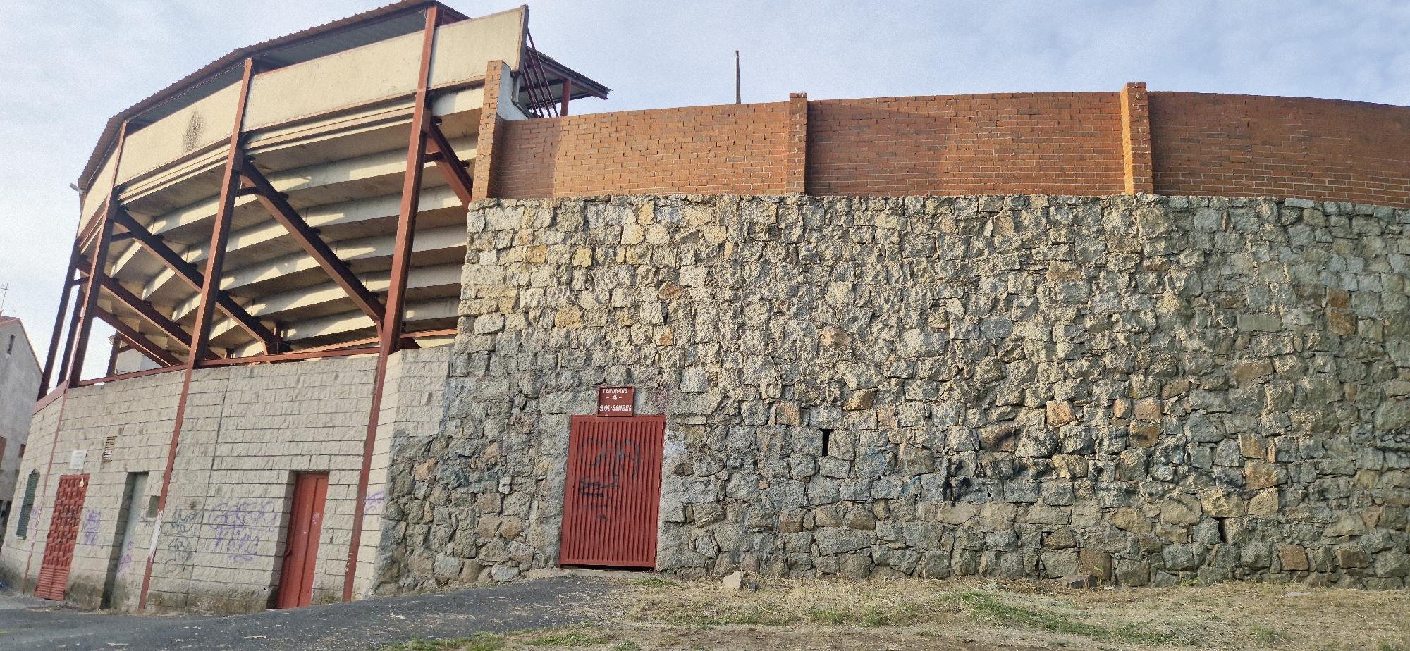 A wide shot captures the exterior of a bullring on a clear day. The structure is composed of several distinct materials. The lower portion of the bullring features a robust, textured wall constructed from rough-hewn stones. On the left side of the image, a section of the bullring is faced with lighter-colored, rectangular brick blocks. Above this, a skeletal framework of reddish-brown metal supports an upper section with tiered concrete seating, visible from the exterior. The uppermost part of the bullring is capped by a curving wall of red bricks. A red metal door with vertical slats is visible centrally on the stone wall, and a smaller red door is present on the lighter brick section to the left. Graffiti is visible on the lighter brick wall near the red doors. The ground in front of the structure is a mixture of dirt and sparse, dry grass. The sky above is a pale blue with a few wispy clouds.