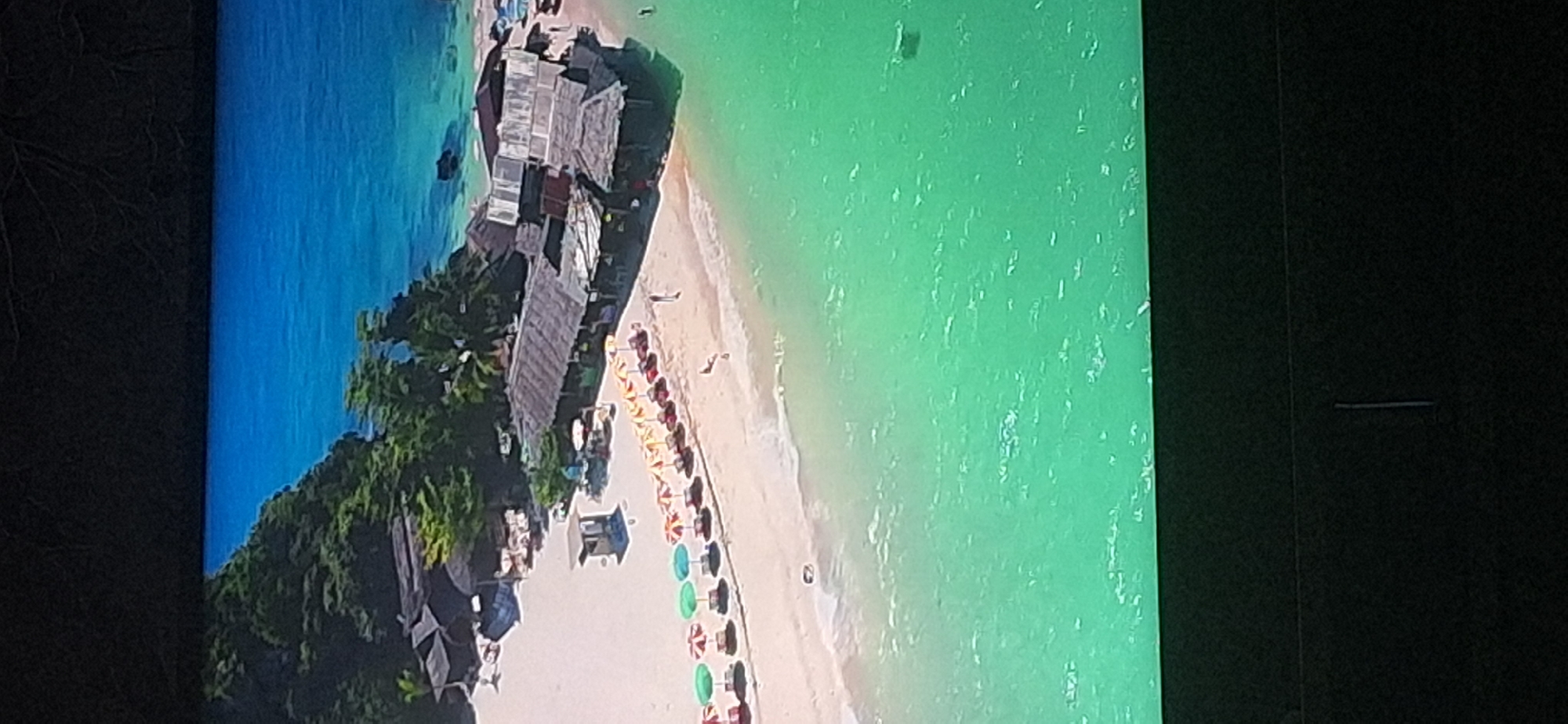 The image shows an aerial view of a tropical beach with turquoise water and white sand. Several thatched-roof buildings are situated on the sand, likely housing shops or restaurants. A row of colorful umbrellas and lounge chairs are set up along the shoreline. Lush green trees and foliage cover a rocky outcrop in the background, with a deep blue ocean extending beyond. There are a few people visible on the beach, including individuals walking and a person in the water. The scene depicts a serene and picturesque beach resort. The location is not Bila Tserkva, Ukraine.