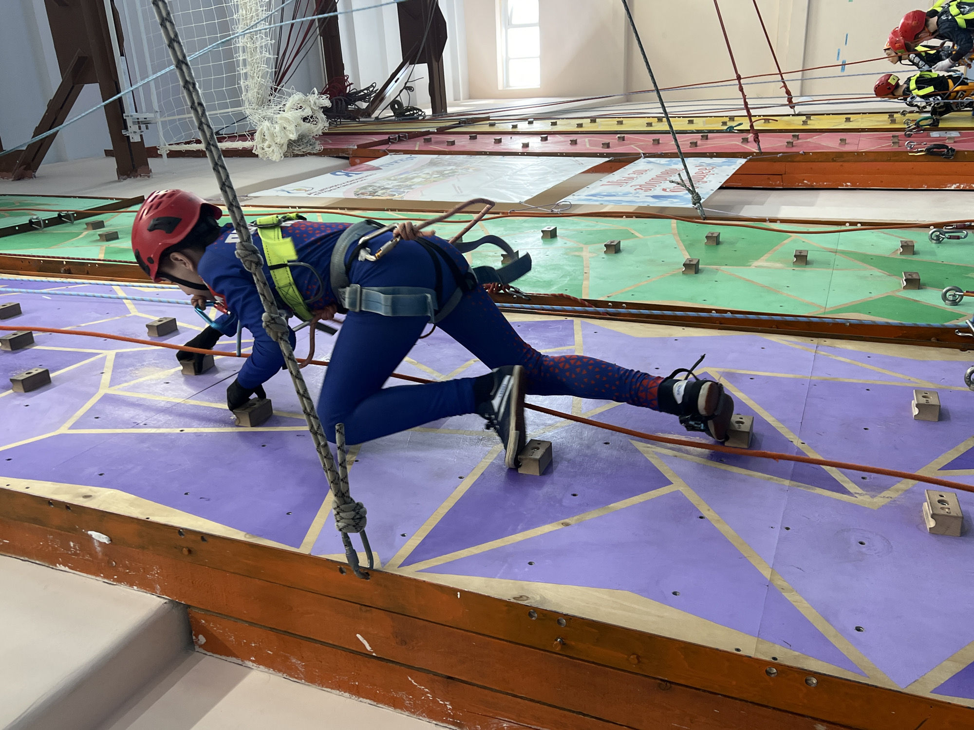A person wearing a red helmet and a blue suit with a climbing harness is traversing a purple climbing wall. The wall is decorated with a geometric pattern of yellow lines and has several rectangular climbing holds attached to it. The person is using a rope for support. The climbing wall is indoors, in a training facility. In the background, there are other climbing walls with different colors and designs. Other people wearing similar gear are also visible on the climbing walls. The facility is located in Berezniki, Russia.