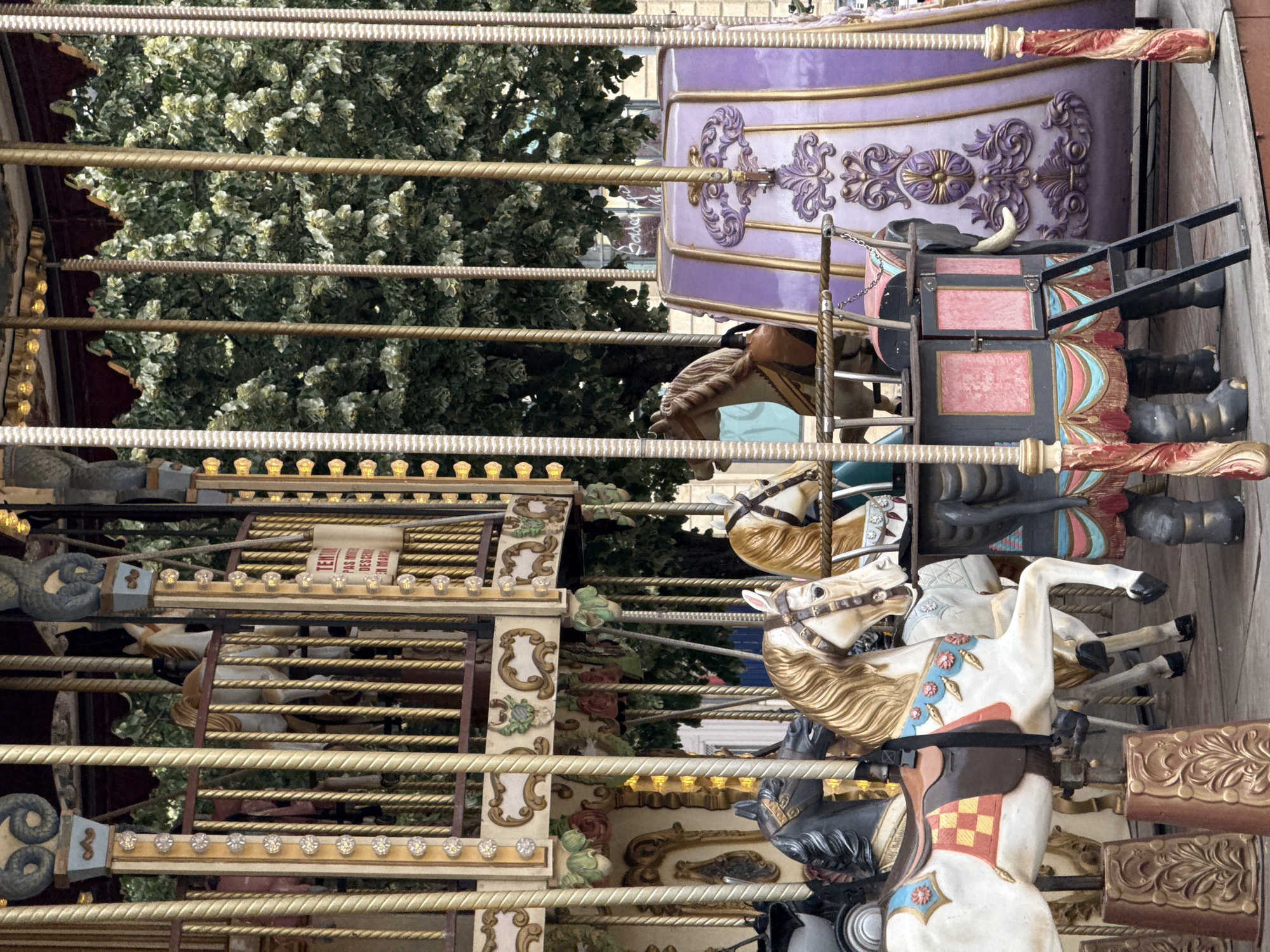 This is a medium shot of a carousel on a cloudy day. The carousel has multiple levels and is adorned with golden poles, ornate carvings, and colorful lights. In the foreground, there are several carousel horses in various colors, including white with gold manes and brown accents, and a black horse. One white horse is prominently featured, with a saddle that has a red and yellow checkered pattern. Behind the horses, there is an elephant figure, part of the carousel's attractions, with a ladder attached to its side. In the background, lush green trees are visible through the carousel's structure. To the right, a purple and gold decorative panel is part of the carousel's design. The carousel is located in the Place de la Bourse in Bordeaux, France, a well-known public square.