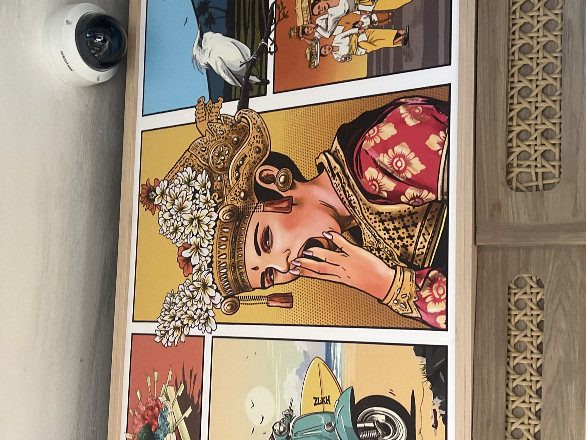 A framed cartoon-style illustration hangs on a wall. The artwork is divided into comic-style panels. These panels depict tropical scenes, including a white bird perched in a tree, people in traditional Indonesian attire, a woman wearing elaborate headwear with flowers and gold adornments, and a blue scooter with a surfboard on a beach. A white security camera is mounted on the wall to the left of the artwork. To the right of the artwork is a vertical strip of woven rattan or similar material. The image was taken in Kuta, Indonesia.