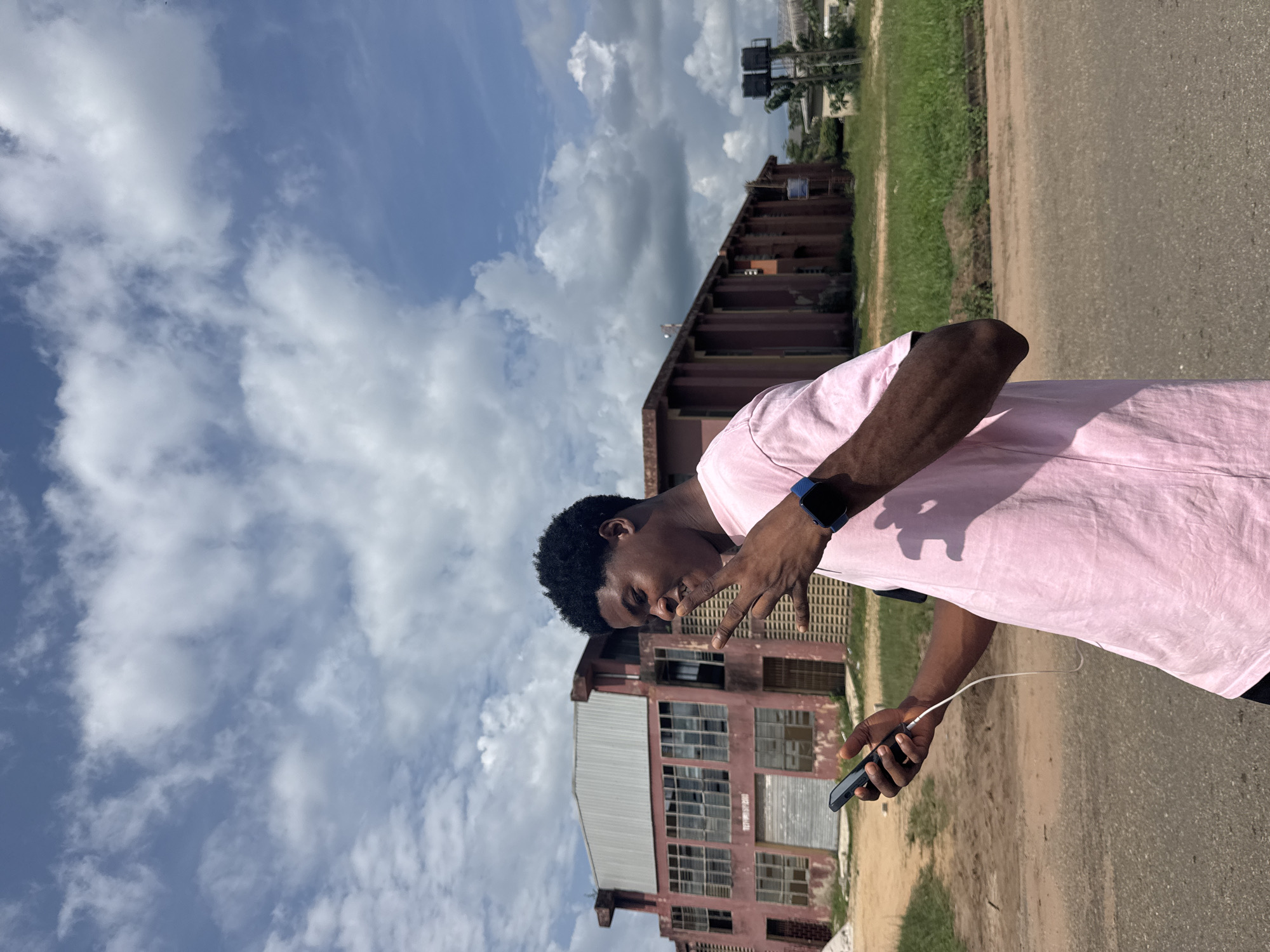 A young man stands outdoors, facing towards the viewer and posing for a photograph. He is wearing a light pink t-shirt and dark pants, with a dark blue smartwatch visible on his left wrist. In his left hand, he holds a dark smartphone, which has a white charging cable connected to it. His right hand is raised, forming a peace sign or V-sign gesture. He has short, dark, curly hair and a relaxed expression.
The scene is set on a bright, sunny day with a clear blue sky scattered with fluffy white clouds, suggesting it is likely mid-morning or afternoon. Behind the man, a large, multi-story building with a reddish-brown facade dominates the background. This building appears somewhat aged or industrial, featuring numerous windows, some of which are broken, boarded up, or reflect the sky. It has a corrugated metal roof. Further in the background, another smaller building, some trees, and utility poles are visible. To the right of the man, a paved road stretches into the distance, bordered by a strip of green grass and dirt. There is faint, illegible text on a sign attached to the large building. The environment suggests an urban or semi-urban area in Ihiagwa, Nigeria.