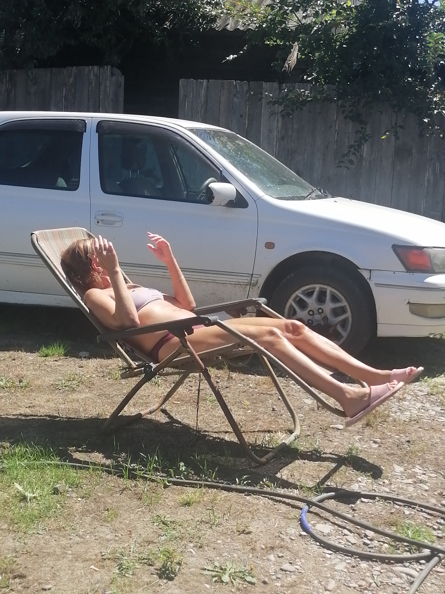 In Novopokrovka, Russia, a woman reclines in a folding chair outdoors, sunbathing. She wears a bikini and appears to be shielding her face from the sun with her hands. A white car is parked behind her, and a wooden fence forms the backdrop. The ground is a mixture of dirt and sparse grass. A garden hose lies coiled on the ground in the foreground.