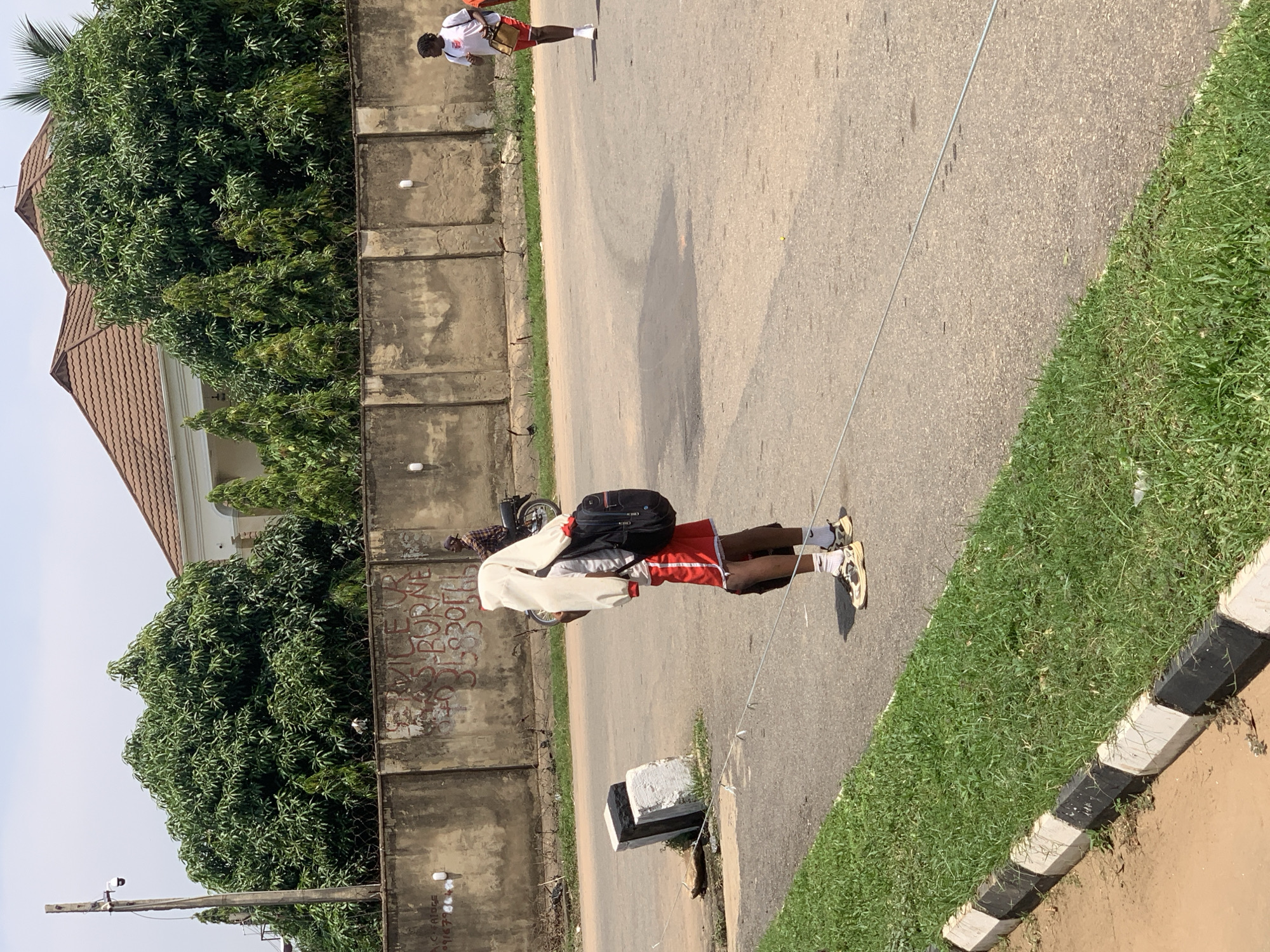 This is a medium-shot, eye-level, outdoor photograph taken on a sunny day, likely in Abuja, Nigeria. The scene depicts a street with a concrete wall in the background, topped with barbed wire and lush green trees behind it. A large, modern house with a brown tiled roof is partially visible above the trees. In the foreground, a young person is standing on the side of the asphalt road, facing away from the camera. They are wearing red shorts, a white t-shirt, and beige sneakers with white socks. A black backpack is slung over their right shoulder, and a white garment is draped over their head and shoulders. Another person, wearing a white t-shirt and red shorts, is walking away from the camera further down the road. In the middle ground, a person on a motorcycle is visible. The road has a white and black striped curb along the grassy edge on the left side of the frame. The overall atmosphere suggests an urban environment with residential and commercial elements.