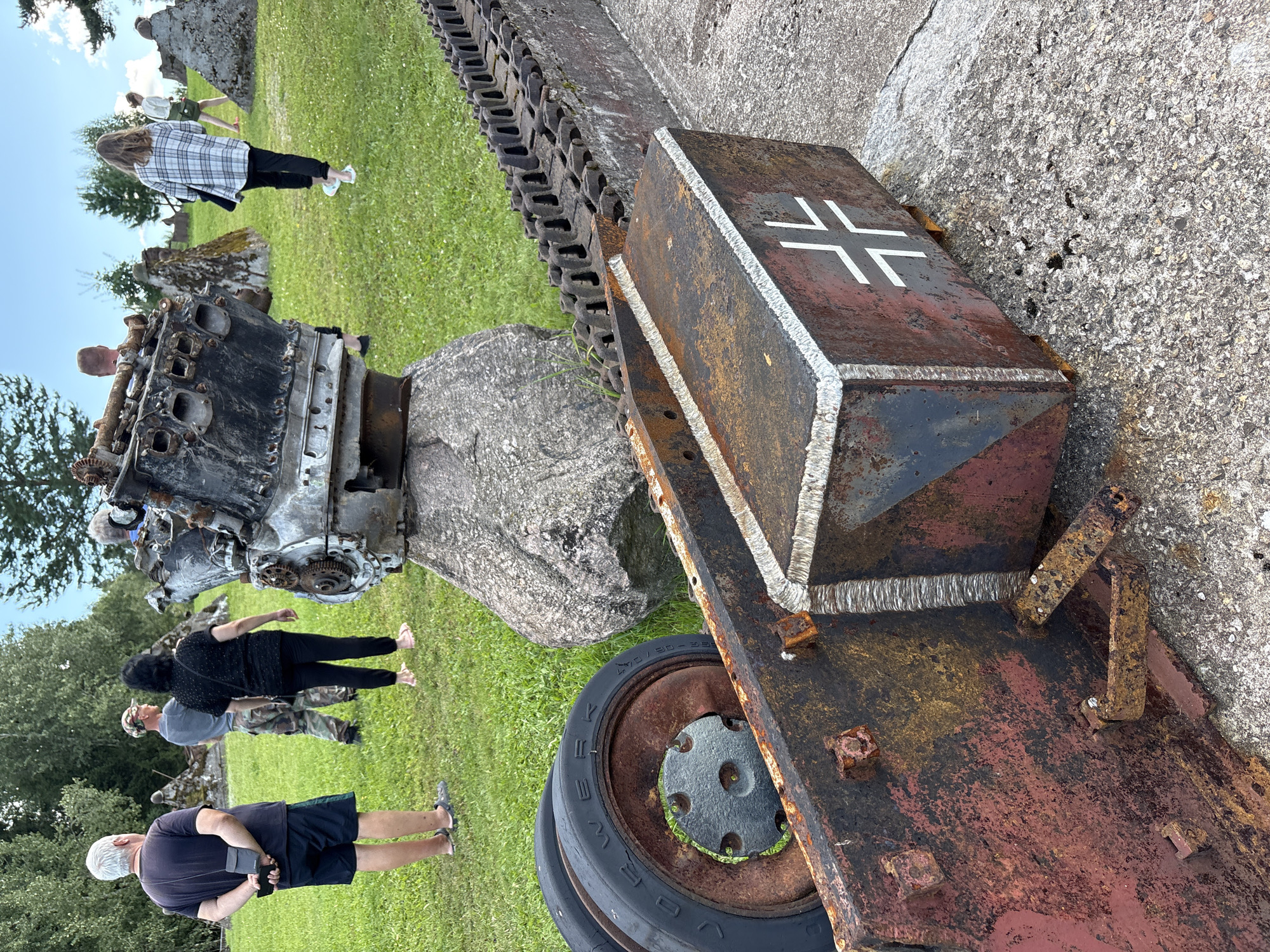 In Zhalybnya, Russia, a group of people are observing remnants of military equipment in an outdoor exhibition. In the foreground, rusted metal components, including a tank track and a wheel with the tire still attached, are visible. A block of rusted metal with a white cross painted on it is also present. In the background, a large, damaged engine block rests on a boulder. Several individuals, some facing away from the camera, are scattered across a grassy area dotted with trees and abstract concrete structures. The sky is partly cloudy.