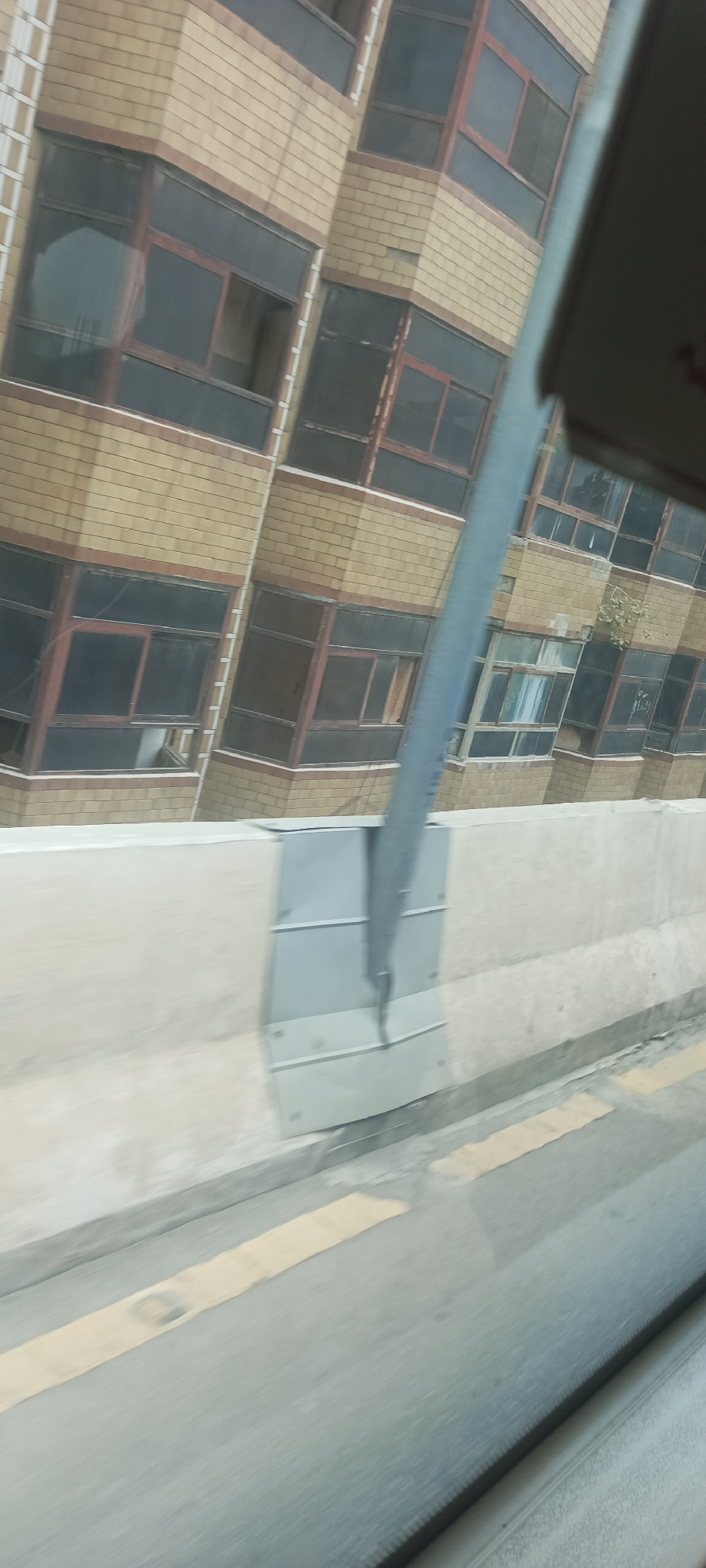 The image captures a scene from a moving vehicle in Alam Khan Kalay, Pakistan. The primary subject is a multi-story building with a distinctive brick facade and numerous windows. The building appears to be an apartment complex or office building, with its architecture exhibiting a modern, yet somewhat weathered, style. The windows are large and dark-tinted, some featuring reddish-brown frames. The building's structure seems to lean or twist, possibly due to perspective or unique architectural design.
In the foreground, a concrete barrier lines the road, with a yellow stripe painted on the asphalt below. A tall, metallic pole, possibly part of a street light or signpost, is prominently positioned in the center of the frame, obscuring parts of the building. The pole is a light gray color and appears to be attached to a metal fixture on the concrete barrier.
The environment suggests an urban or semi-urban setting. The lighting indicates daytime, with overcast skies contributing to a diffused light. There are no people clearly visible in the image, and the focus is on the architecture and infrastructure. The image is slightly blurred, indicating motion, as if taken from a car or bus window.