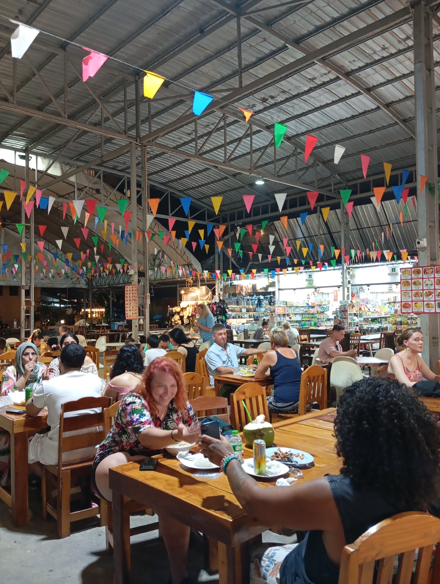 The image depicts an outdoor dining area in Karon, Thailand, likely a night market or open-air restaurant. Numerous colorful triangular flags are strung across the metal ceiling structure, creating a festive atmosphere. Several tables with wooden chairs are occupied by a diverse group of people dining and socializing.
In the foreground, a woman with curly black hair is seated at a wooden table, facing away from the camera. She is wearing a black sleeveless top and appears to be interacting with a smartphone in her hand. On the table in front of her are a plate of food, a can of soda, and a halved coconut with a straw. To her left, a woman with red hair and a floral shirt is smiling and looking towards the camera. She has a plate of food and a drink on her table.
Other diners are visible at various tables throughout the scene. Some are engaged in conversation, while others are eating or looking at their phones. The background shows rows of shelves stocked with goods, suggesting the presence of a market or shops adjacent to the dining area. The lighting is dim, indicative of an evening setting. The overall impression is one of a lively and casual dining experience.