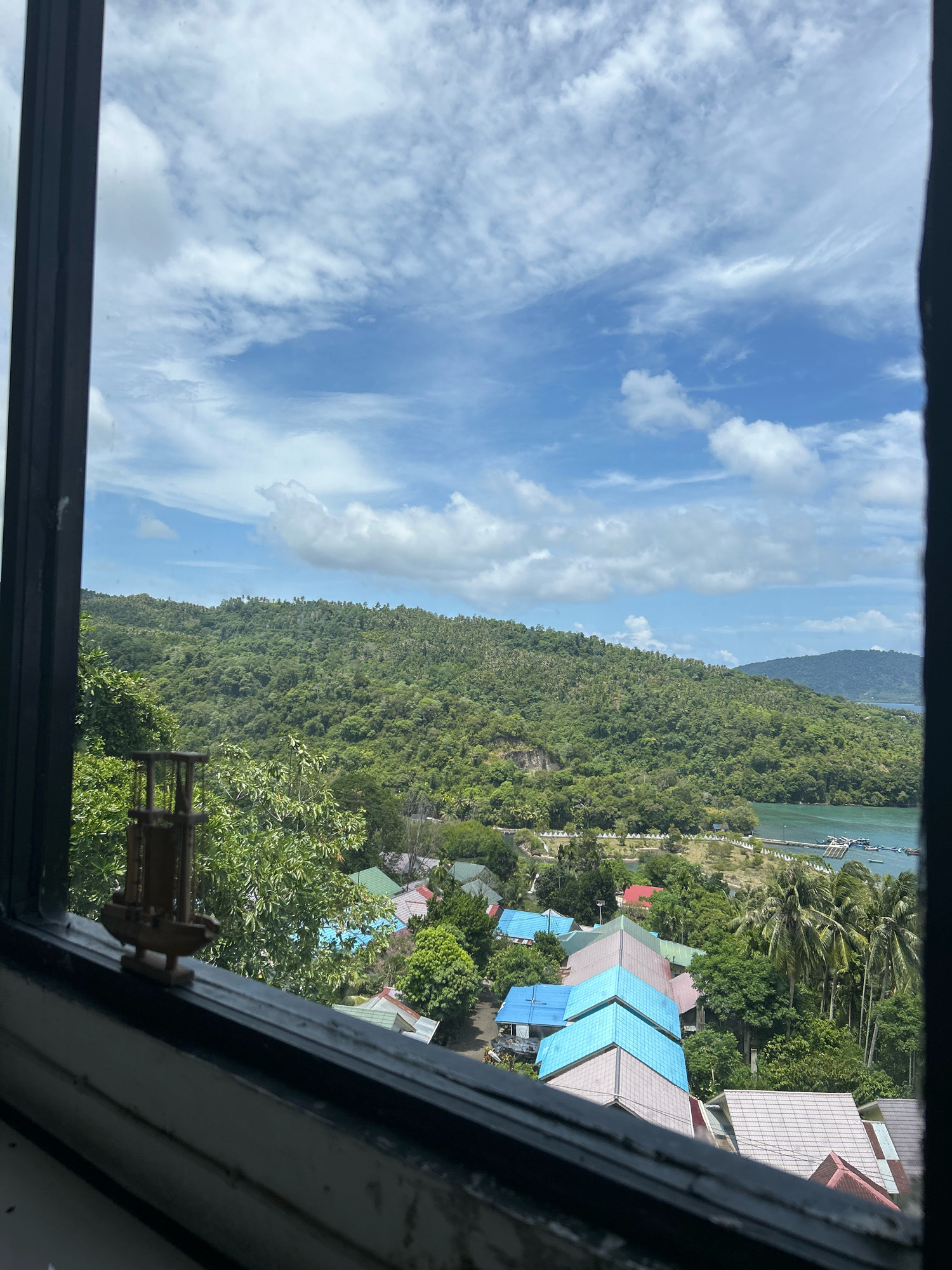 The image, taken from a window, shows a view of Sabang, Indonesia, with a lush green hillside dominating the background. The hillside is covered in dense tropical vegetation, with a few scattered palm trees visible. A body of water, likely the sea, is visible to the right of the hillside, with a pier and several boats. In the foreground, a cluster of buildings with brightly colored roofs – blue, pink, and gray – are nestled amongst trees. A small, wooden model of a sailboat sits on the windowsill in the lower left corner of the frame. The sky above is a vibrant blue, dotted with fluffy white clouds. The scene suggests a tropical coastal environment.