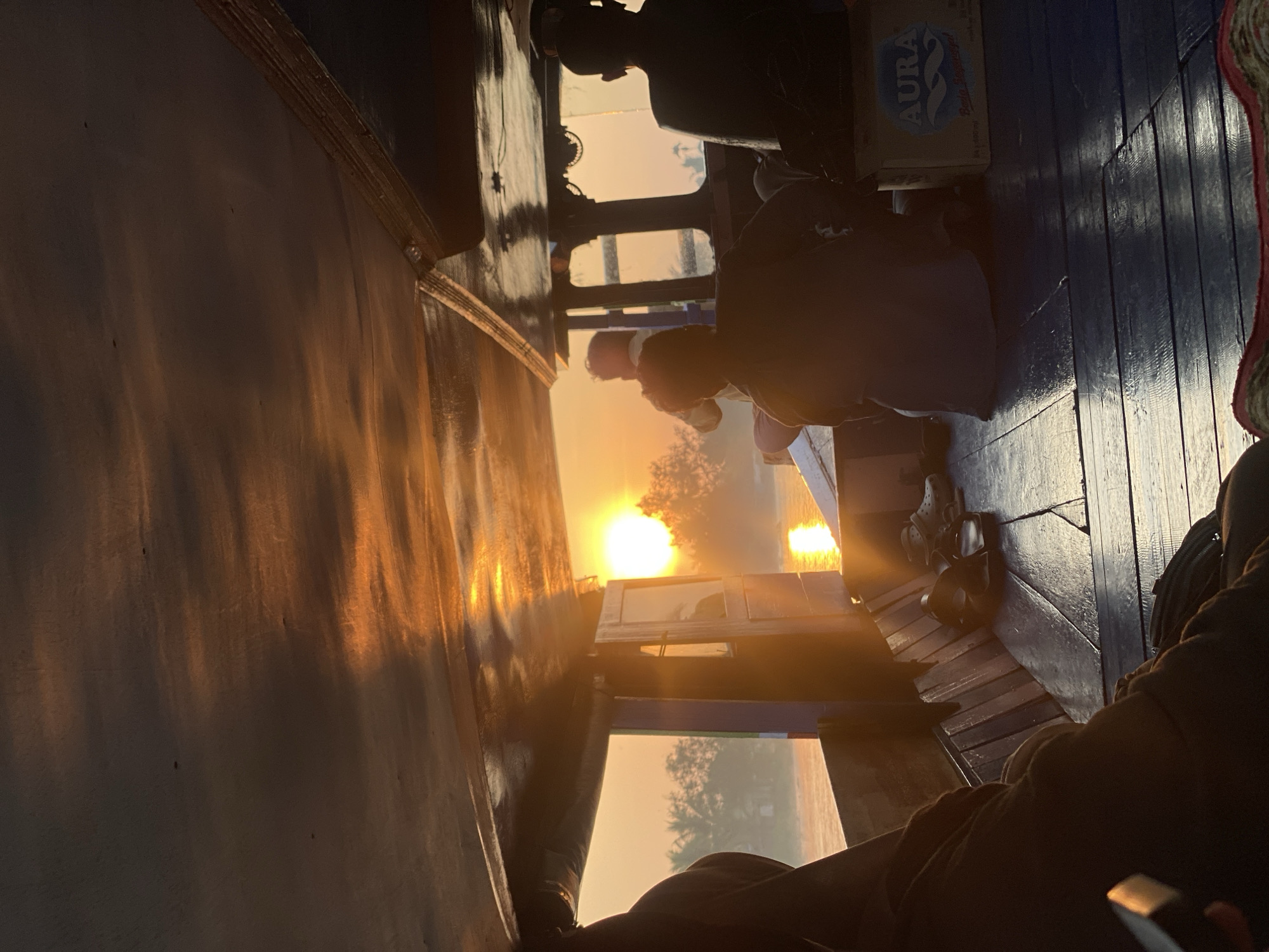 The image captures a serene sunrise scene from inside a wooden boat navigating the waters of Sungai Tandipah, Indonesia. The low angle shot looks towards the front of the boat, with the underside of the roof of the cabin illuminated by the warm glow of the rising sun. The sun itself is a bright, distinct orb low on the horizon, casting a golden sheen across the water and reflecting off the wet wooden deck of the boat.
Several people are visible within the boat, silhouetted against the bright light. In the foreground, on the left, a person in a dark hooded garment is seated, with their back to the camera. To their right, a pair of dark sandals and a grey shoe are placed on the deck. Further into the boat, in the center, another person is seated, facing away from the camera, wearing a dark blue garment. To their right, a person in a dark shirt is also seated, with their back to the camera. On the far right, a box labeled "AURA" is visible, indicating it contains bottled water.
The environment outside the boat shows lush greenery, with palm trees lining the riverbank, suggesting a tropical setting. The water appears calm, with ripples reflecting the sunlight. The overall atmosphere is peaceful and tranquil, characteristic of a sunrise boat journey in a natural landscape. The wooden interior of the boat, with its worn planks and visible grain, adds to the rustic charm of the scene.