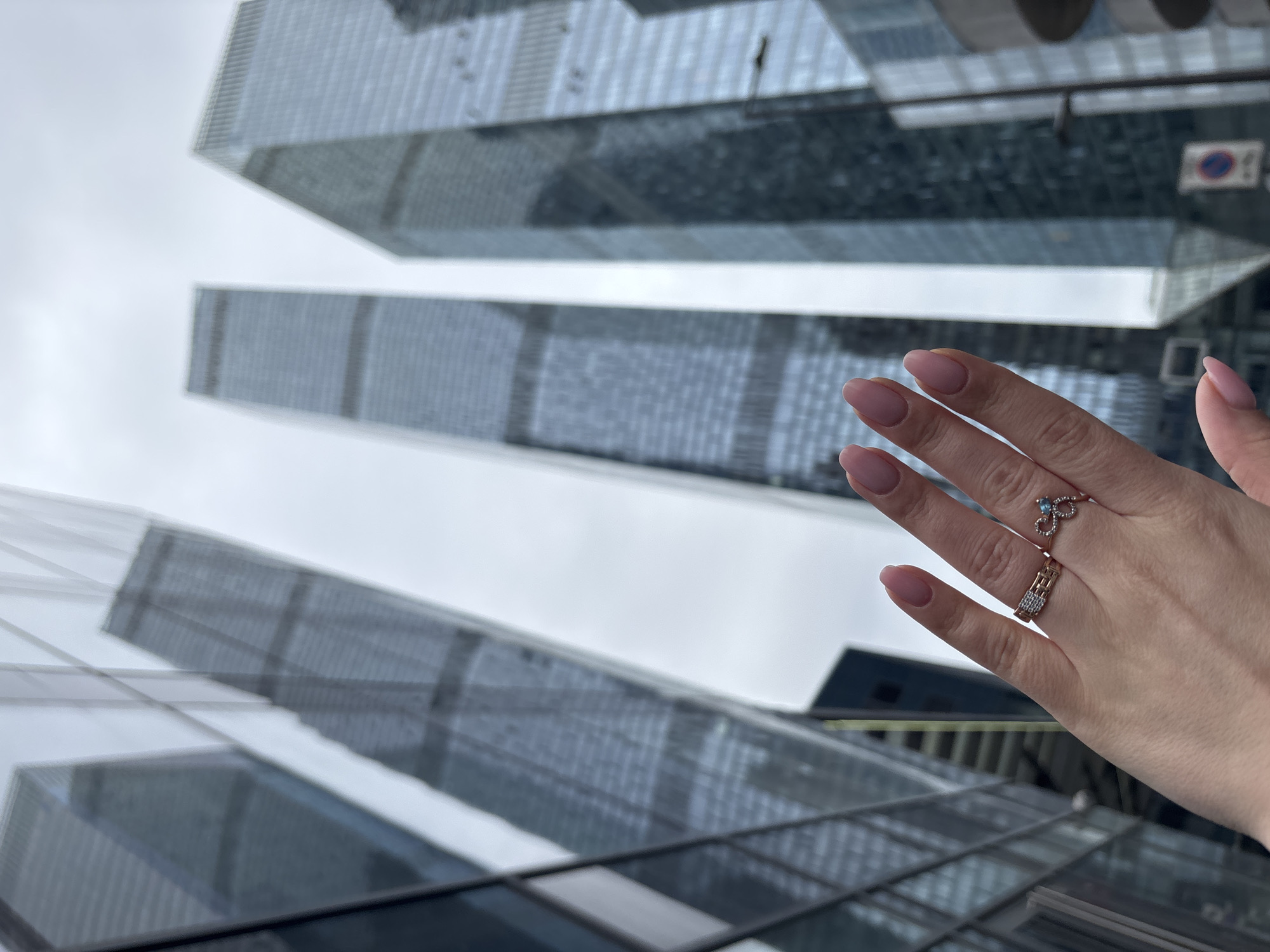A low-angle shot captures several skyscrapers, seemingly in Moscow, Russia, against a cloudy sky. In the foreground, a fair-skinned hand with manicured nails is visible. The hand features two rings: one with a geometric design and another with a small blue stone. The skyscrapers are modern, with glass facades reflecting the overcast sky. The angle suggests the photo was taken from a low vantage point, possibly street level. A small sign with a blue circle and red line through it is visible near the top right of the frame.