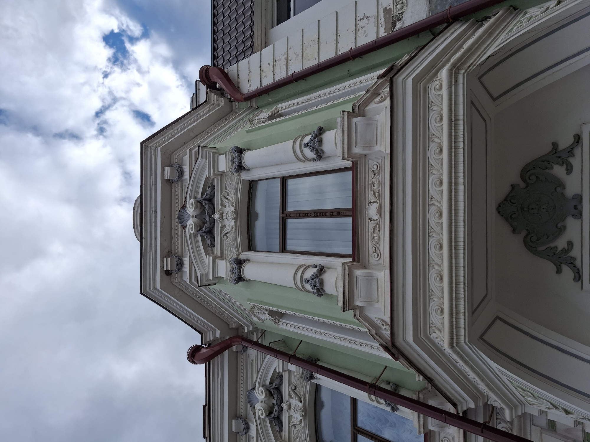 This low-angle photograph captures the ornate facade of a historic building in Sarapul, Russia, against a cloudy sky with glimpses of blue. The architecture is characterized by elaborate, off-white stucco detailing that frames a central section painted a pale green.
The building features intricate decorative elements, including classical cornices, moldings, columns with ornate capitals, and scrolled volutes. Around the windows, there are decorative clusters, possibly representing fruits or grapes, adding to the rich ornamentation. Two windows with dark brown frames are visible, reflecting the sky. Above the green section and running across the facade, a reddish-brown downspout is prominently featured, hinting at the building's infrastructure.
At the very top, dark, possibly tiled, roofing is visible. The facade shows signs of age and weathering, with minor discoloration and some peeling, indicating its historical character. The image is taken during the daytime, under an overcast sky. No people or visible text are present. The overall impression is one of detailed craftsmanship and grand, classical architectural styling.