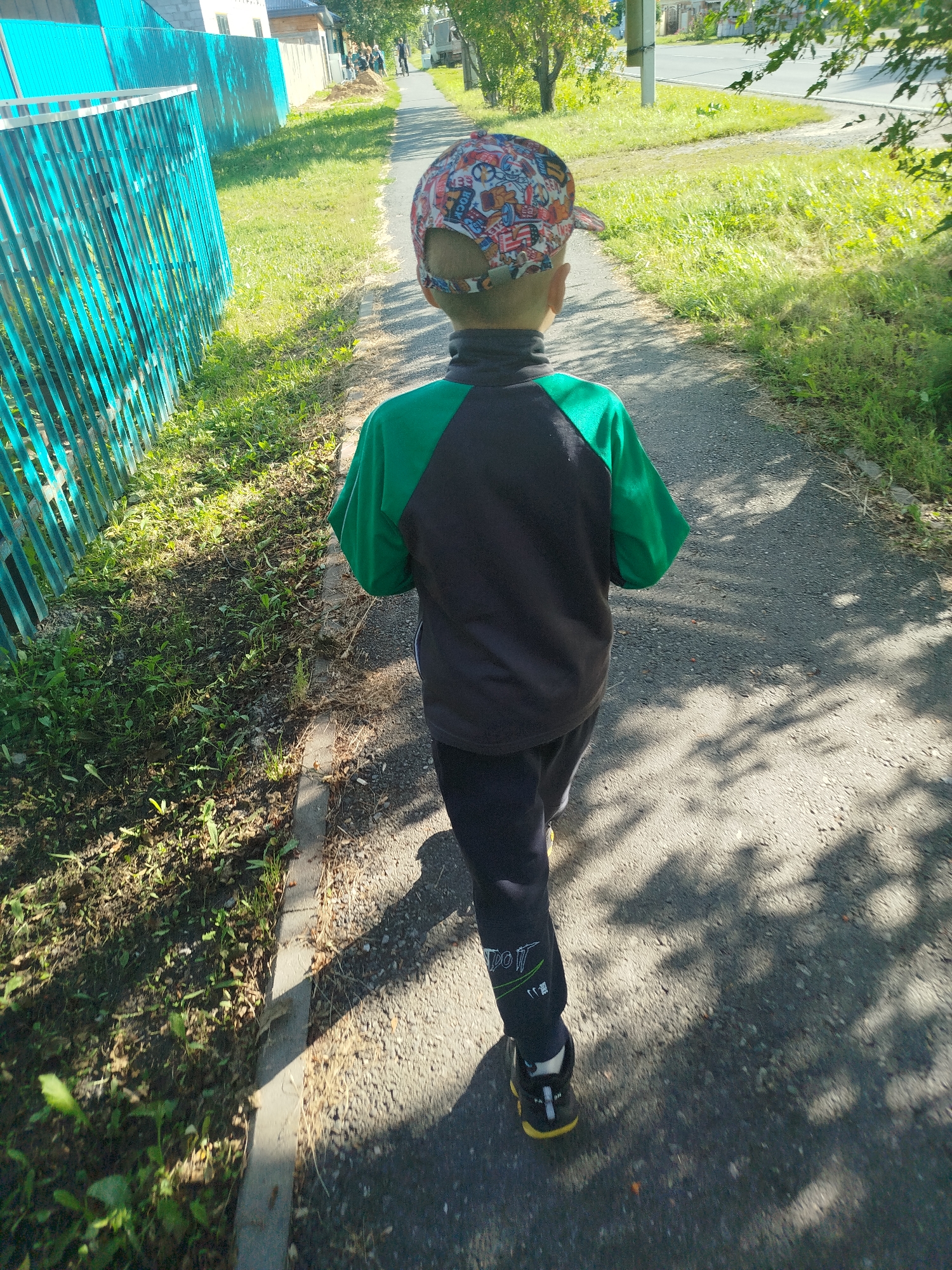 A young boy wearing a colorful baseball cap and a green and black jacket is walking away from the camera down a paved path. The path is lined with a turquoise metal fence on the left and green grass and trees on the right. In the background, houses and a street are visible, suggesting an urban or suburban setting. The lighting indicates it is daytime, likely morning or afternoon, with dappled shadows from the trees falling across the path. The image was captured in Zavodoukovsk, Russia.