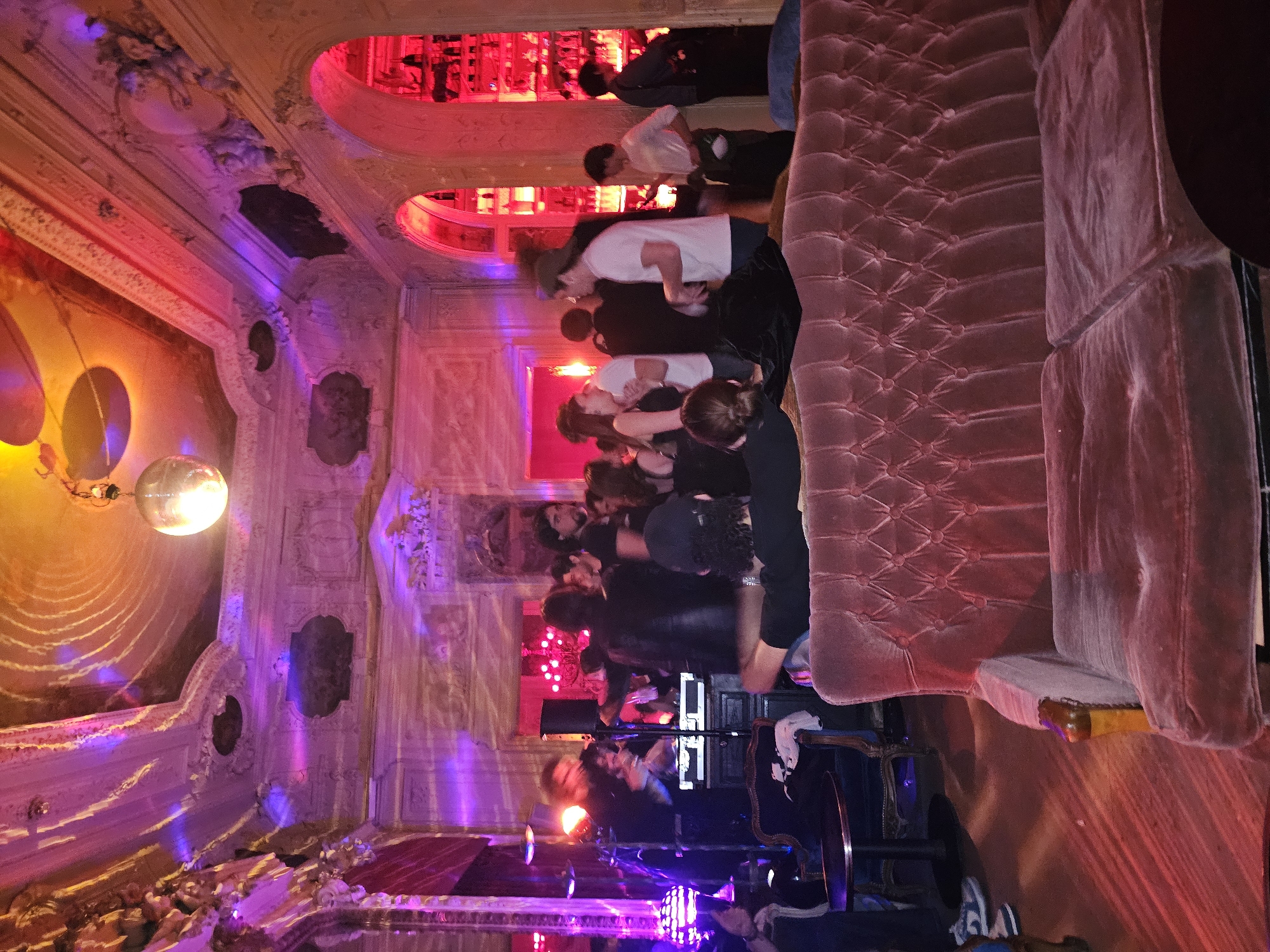 A dimly lit Parisian interior showcases a group of individuals gathered near a velvet tufted sofa, possibly in a lounge or club setting. The ornate architecture of the room features decorative molding, rounded openings, and colorfully lit niches with vibrant hues of pink, purple, and orange illuminating the scene. A disco ball hangs from the ceiling, casting speckled light patterns. People, including a band performing, are positioned in front of musical equipment.