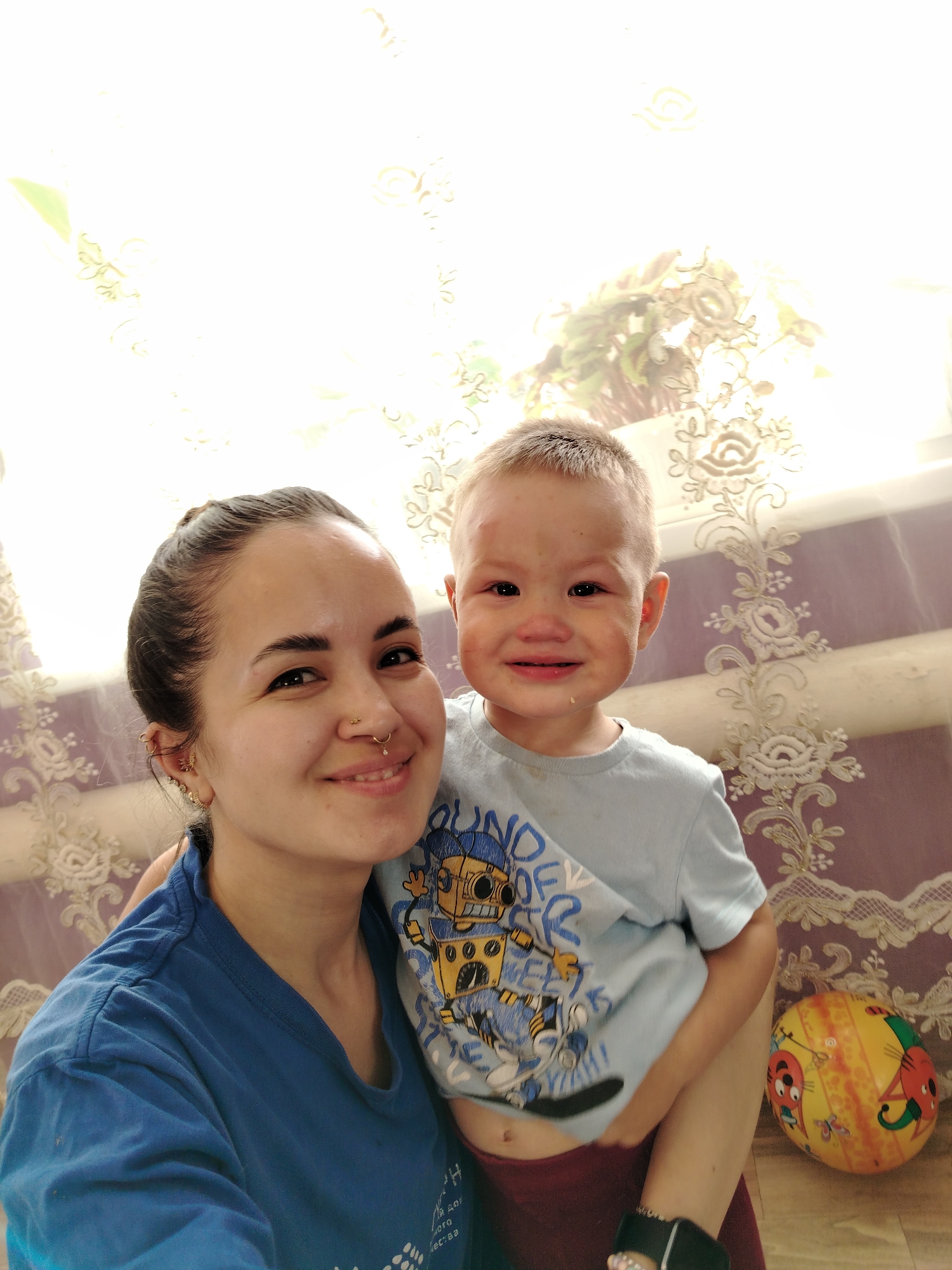 A selfie-style shot features a woman and a young child indoors, likely in a home environment. The woman, with dark hair pulled back in a bun, wears a blue t-shirt and multiple piercings in her ear and nose. She smiles at the camera with her arm around the child.
The child, fair-skinned with light hair, wears a blue t-shirt with a cartoon robot design. Despite appearing tearful, with visible wetness on his face, he seems to be trying to smile.
Behind them, a window covered with patterned lace curtains provides soft, diffused lighting. The backdrop is a wall covered in a purple and beige floral wallpaper. A brightly colored, cartoon-decorated ball sits on the floor to the right.
The image was taken in the city of Гожан, Russia.