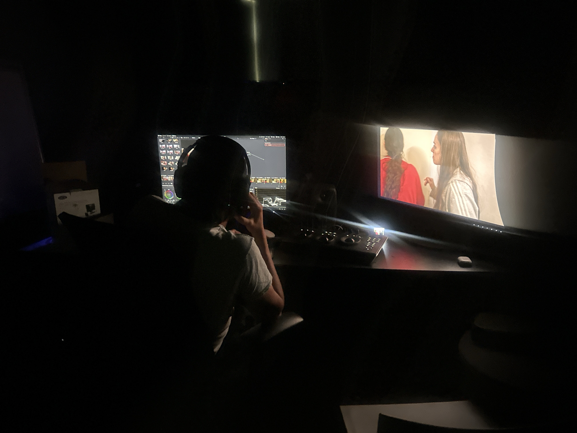 A person wearing headphones sits in front of two computer monitors in a dark room. The monitor on the left displays editing software, while the one on the right shows a scene with two women, one wearing a red shirt and the other a white shirt. The person appears to be working, with a hand raised towards their face as if in thought or contemplation. A control panel with knobs and buttons sits on the desk in front of them. Additional objects and furniture are dimly visible in the foreground and background. The location is not discernible from the image.