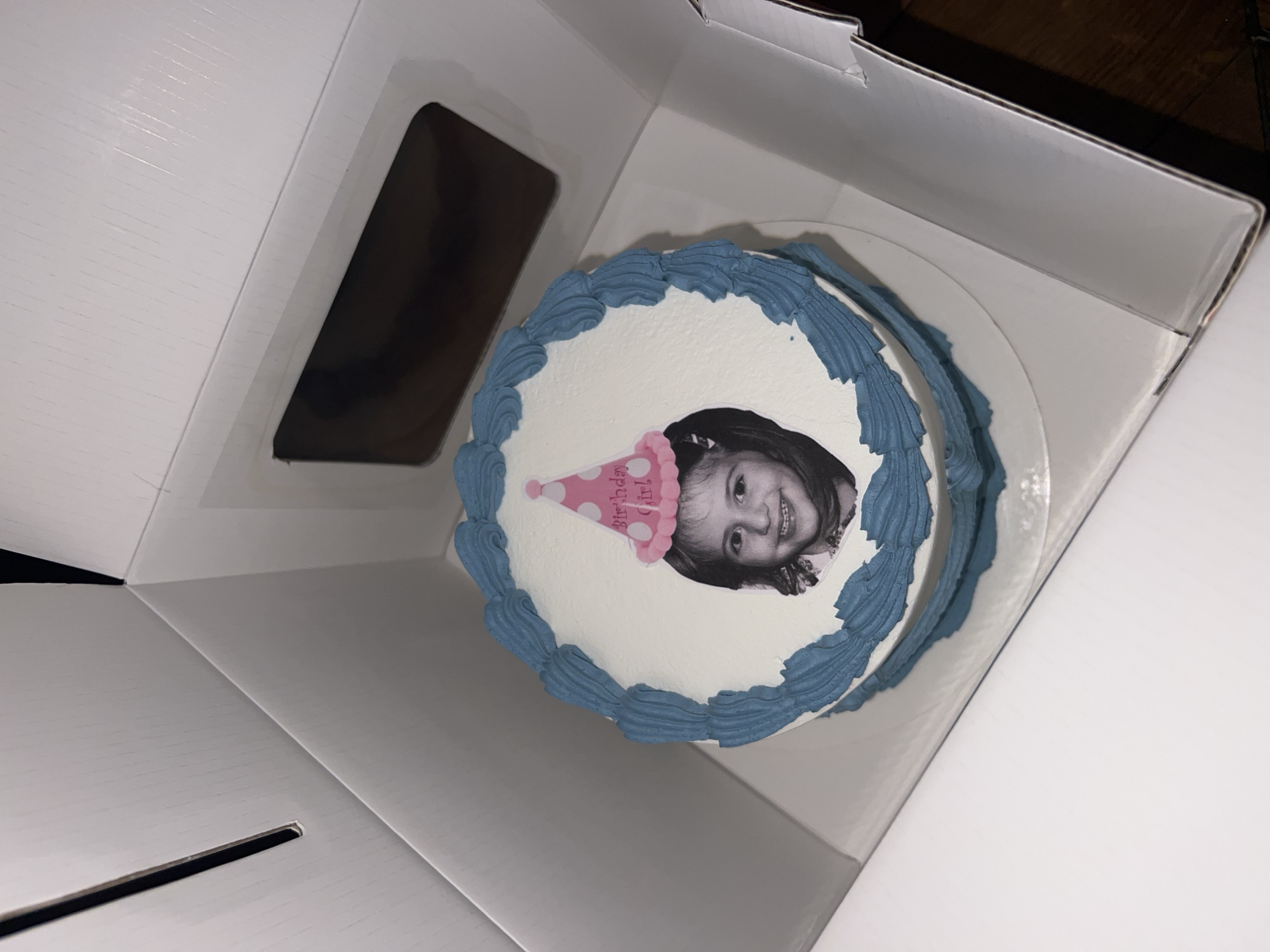 The image shows a cake in a white cardboard box. The cake is round, with white frosting and a blue decorative border. A black and white photograph of a young girl wearing a party hat is centered on the cake. The box has a rectangular cutout on one side. The scene is indoors, and the lighting is somewhat dim. The surrounding environment is not clearly visible. This is not a scene in Paris, France.