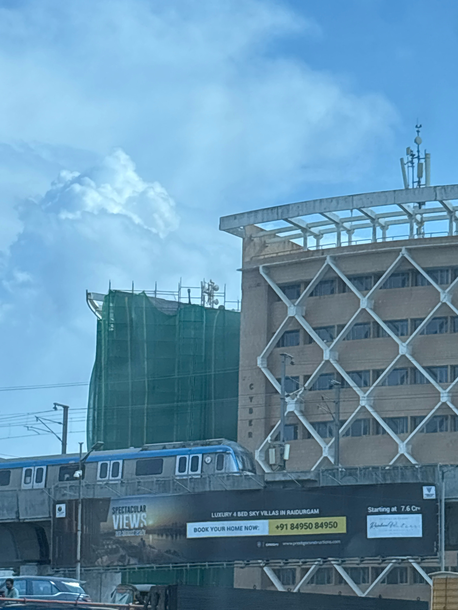 The image depicts an outdoor urban scene with a clear blue sky and scattered clouds. In the foreground, a train is visible on elevated tracks, with a large billboard below it advertising "SPECTACULAR VIEWS LUXURY 4 BED SKY VILLAS IN RAIDURGAM" and a phone number "+91 84950 84950". The billboard also indicates a starting price of "7.6 Cr". To the right of the train and billboard is a multi-story building with a distinctive diamond-shaped facade pattern. Behind this building, a partially constructed structure is covered in green netting. Communication antennas are present on top of some of the buildings. The overall impression is a busy urban environment with ongoing construction and modern infrastructure. Based on the billboard mentioning "RAIDURGAM", the location is likely Raidurgam, a sub-region in Hyderabad, India, known for its commercial and residential developments.