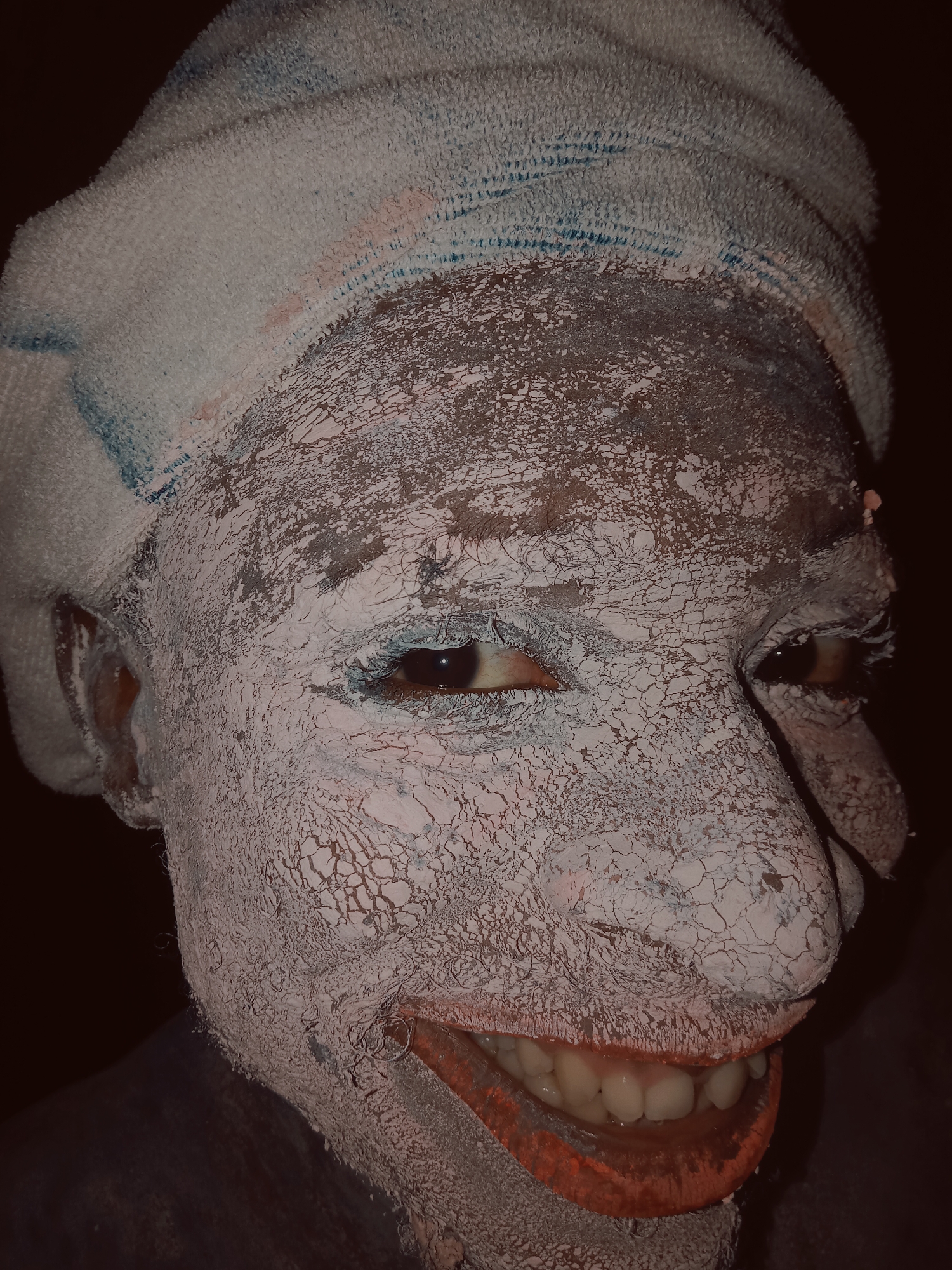 A close-up portrait features a person whose face, ears, and visible neck are entirely covered in a thick, cracked white substance, resembling dried clay or chalk, set against a dark, indistinct background. A white, terry cloth towel, marked with blue and faint pinkish stripes or stains, is wrapped around their head.
The person is captured in a wide, open-mouthed smile, revealing a set of white teeth and pink gums. Their lips are painted with a vibrant red or orange pigment, contrasting sharply with the white face covering. Their brown eyes are visible through the cracked substance, accented by what appears to be light blue pigment around the eyelids. The white substance on the face displays an intricate network of cracks, creating a textured surface.
The image conveys an emotion of joy or amusement through the clear smile. The overall presentation, including the face paint and headwrap, suggests a ceremonial, festive, or performance context, aligning with cultural practices often found in places like Onitsha, Nigeria. The lighting is low, with the face being the primary illuminated subject. There is no visible text.