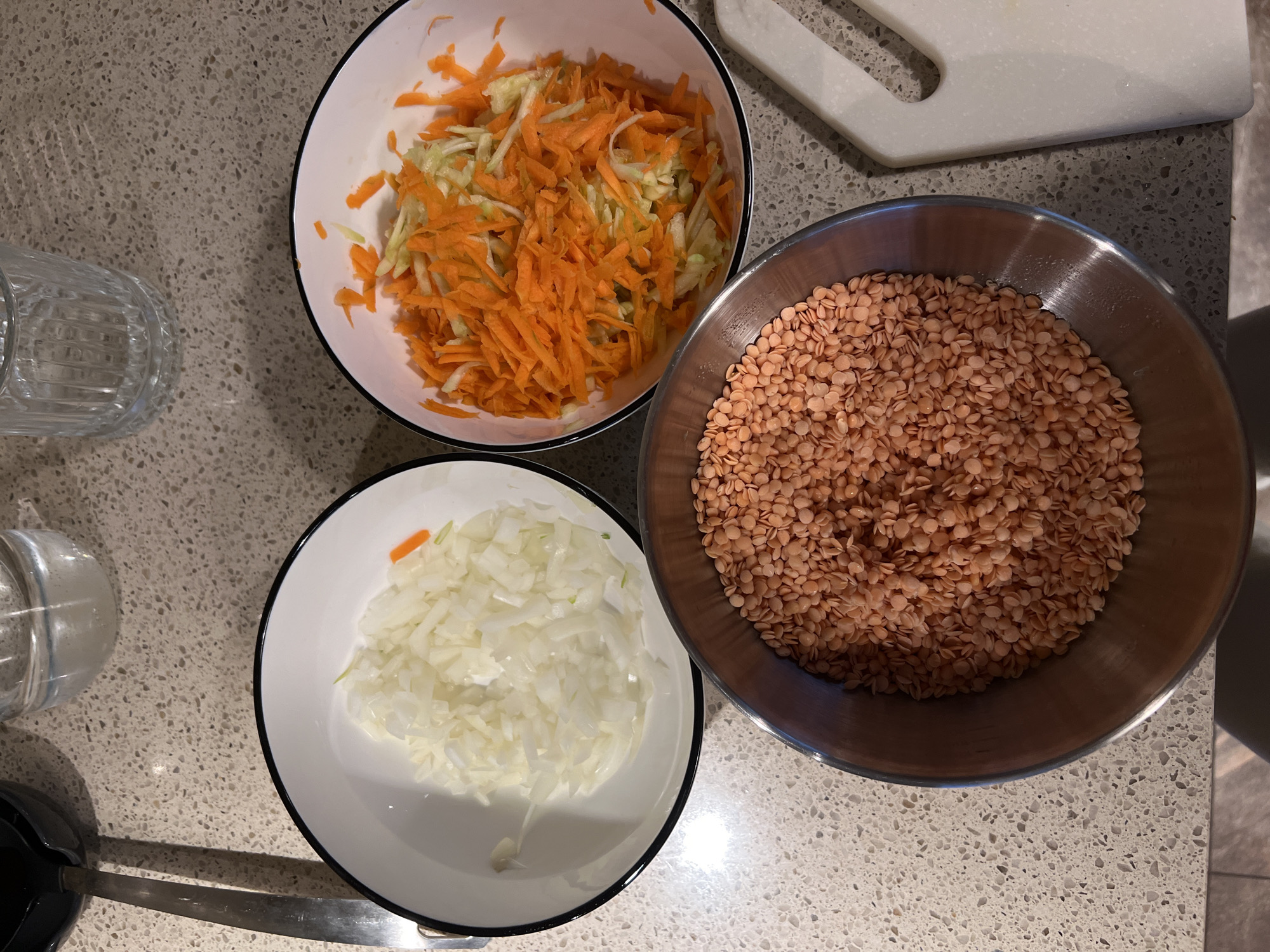 The image shows a preparation scene, likely for cooking. There are three bowls on a speckled countertop. The bowl at the top contains shredded carrots and cabbage. The bowl on the lower-left contains diced white onion. The bowl on the right, which is stainless steel, is filled with red lentils. A glass, ladle, and cutting board are partially visible around the bowls. The location is not evident in the image, and there is no indication that the image was taken in Khimki, Russia. No people are visible. The subject is food preparation.