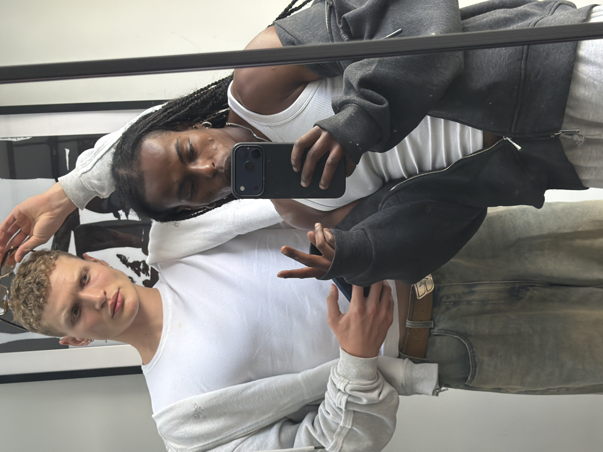 The image captures a mirror selfie of two individuals in what appears to be a domestic setting, possibly an apartment in London, United Kingdom. The person on the left, with fair skin and short, curly blonde hair, is wearing a white ribbed t-shirt and a grey hoodie draped over his shoulders. He is holding a pair of sunglasses above his head and looking towards the camera. The person on the right, with dark skin and long, braided hair, is wearing a white ribbed tank top and a dark grey zip-up hoodie. He is holding a smartphone in his right hand, actively taking the photo, and his left hand is extended, making a peace sign. The background is simple, with a large framed artwork on the wall and a white wall. The clothing suggests a casual, relaxed atmosphere. The overall lighting is bright, with no clear indication of time of day.
